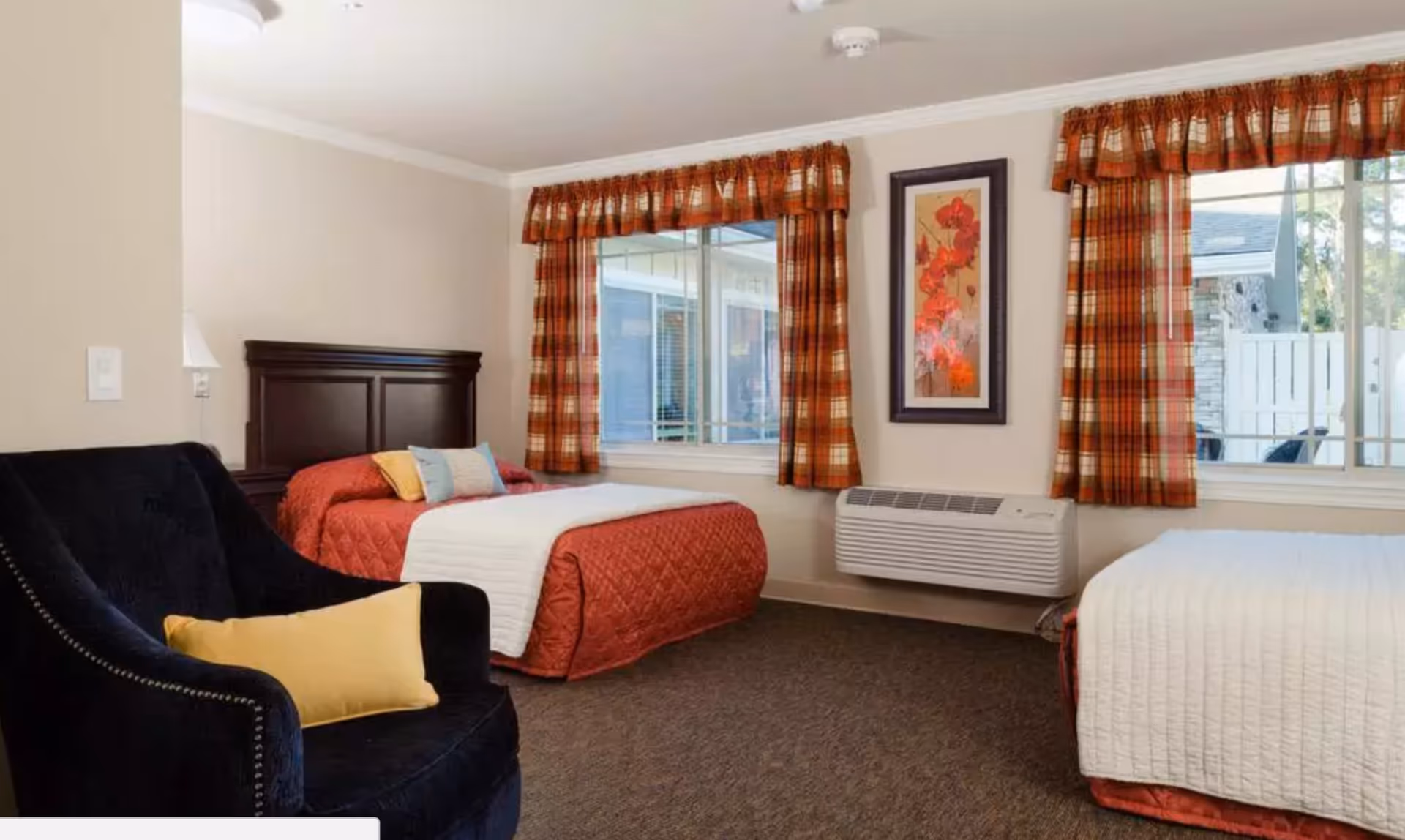 A cozy bedroom with two beds, one with a red quilt and the other with a white quilt. Both beds have pillows and are positioned near two windows with plaid curtains. A dark upholstered armchair with a yellow pillow is in the foreground. A framed floral artwork hangs on the wall above a wall-mounted air conditioning unit.