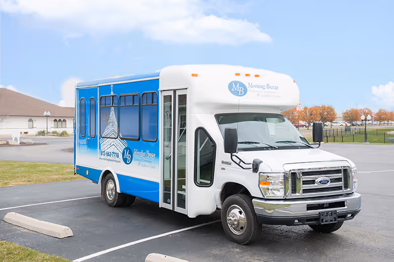 A white and blue shuttle bus parked in a parking lot near a building. The bus has the logo and name 'Morning Breeze Retirement Community & Healthcare Center' on the front and side. The background shows a partly cloudy sky, some trees with autumn foliage, and a grassy area.