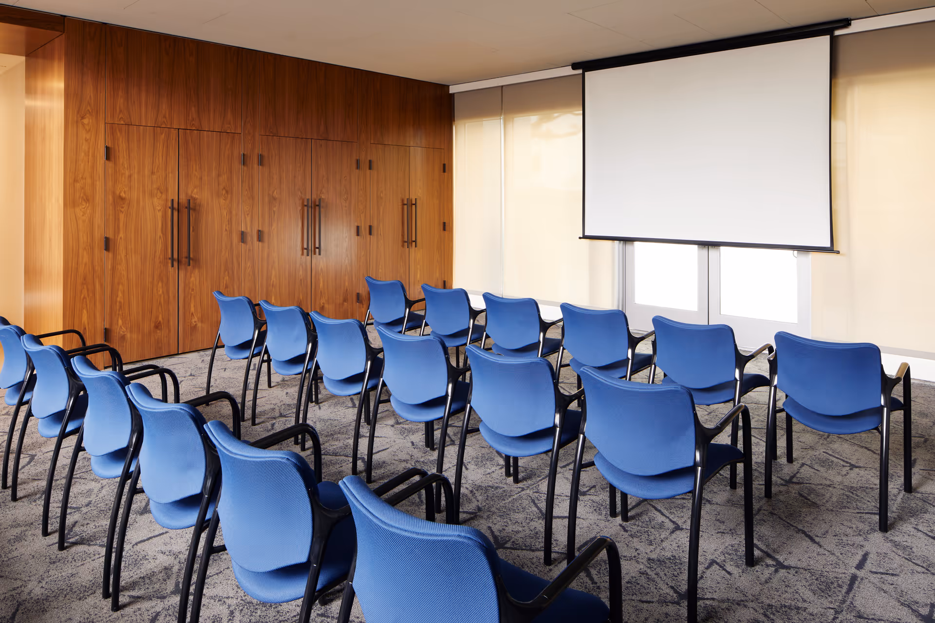 A small conference or presentation room with rows of blue chairs facing a blank white projection screen. The room has wood-paneled walls on one side and large windows covered with beige roller shades on the other side.