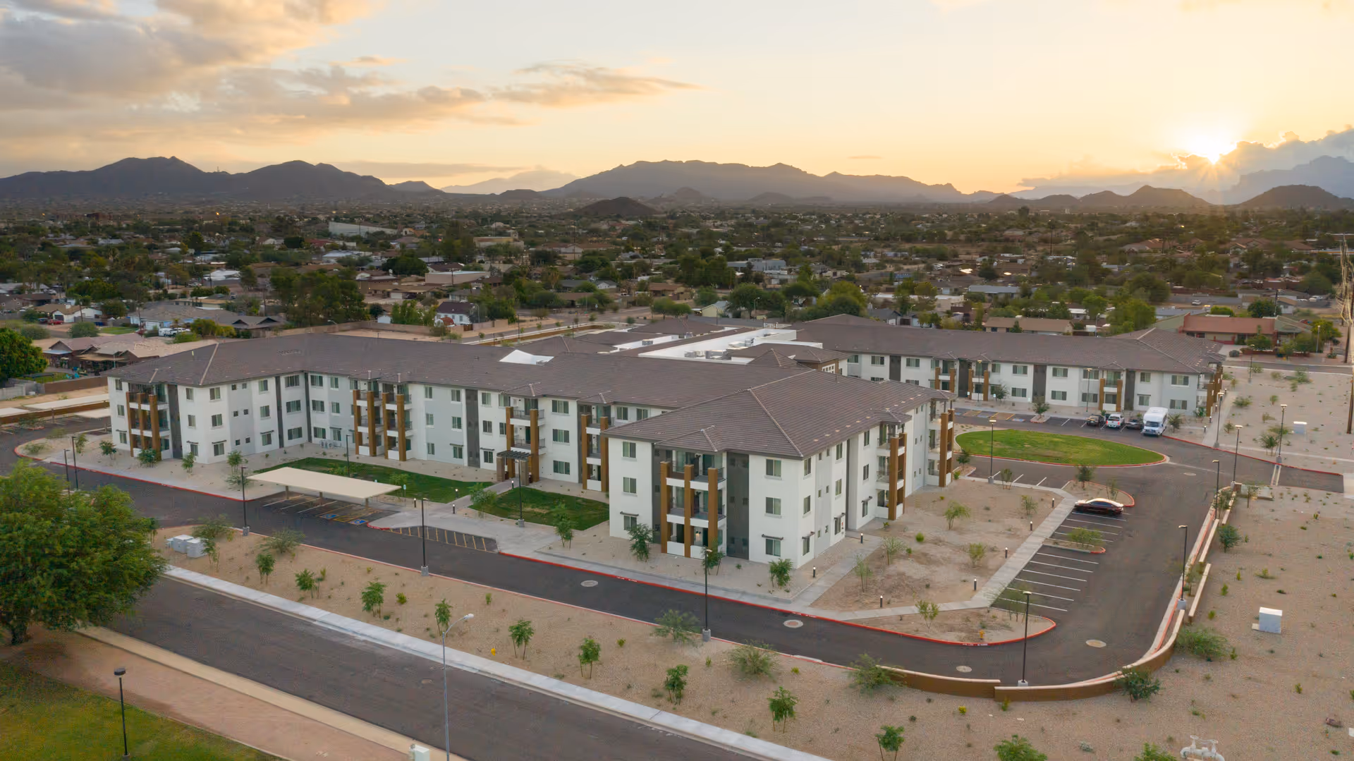 Aerial view of a large, modern three-story senior living facility named ACOYA Mesa, surrounded by desert landscaping and parking areas, with mountains in the background under a sunset sky.