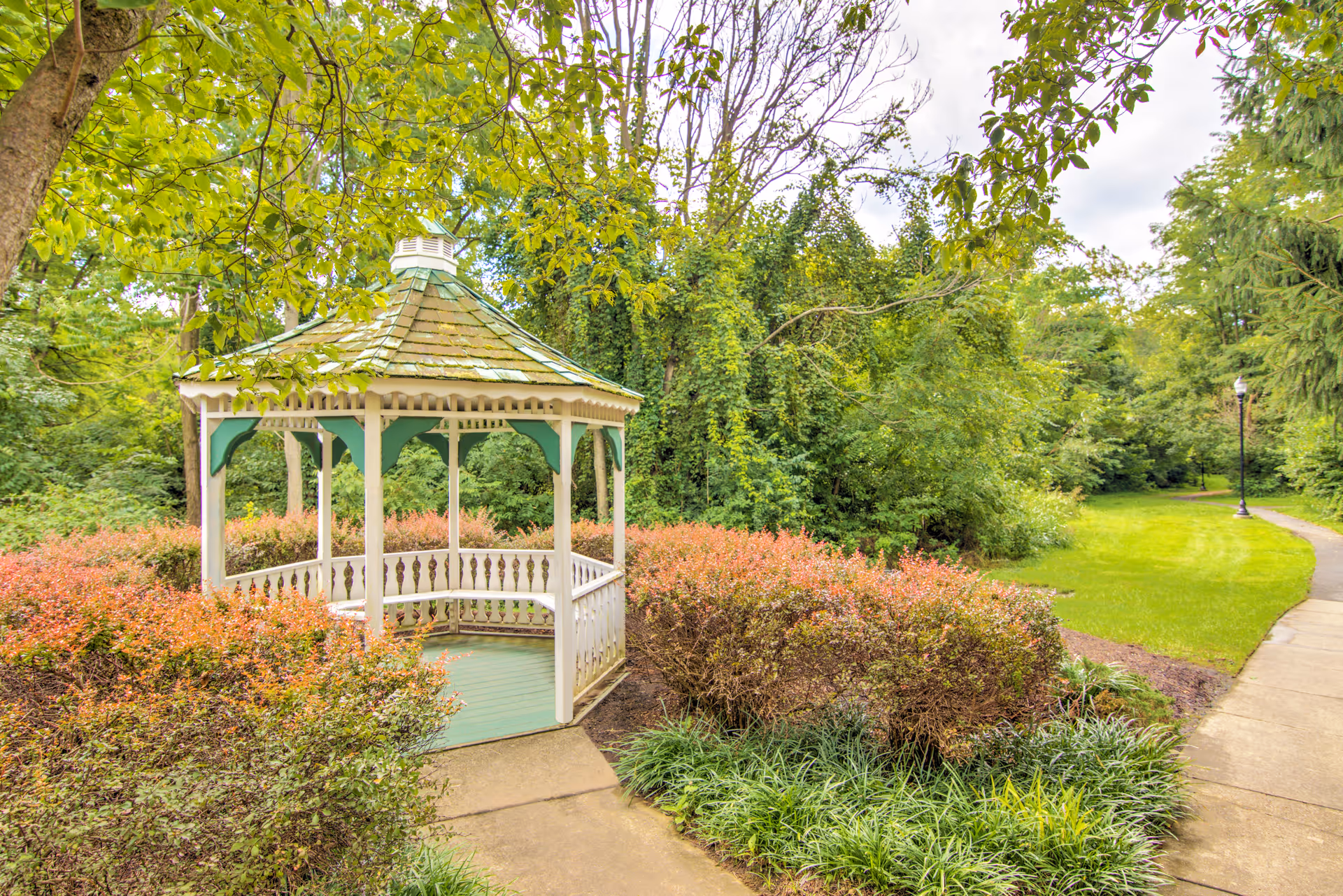 A white and green wooden gazebo surrounded by bushes and trees in a lush garden area with a paved walkway curving to the right, under a partly cloudy sky.