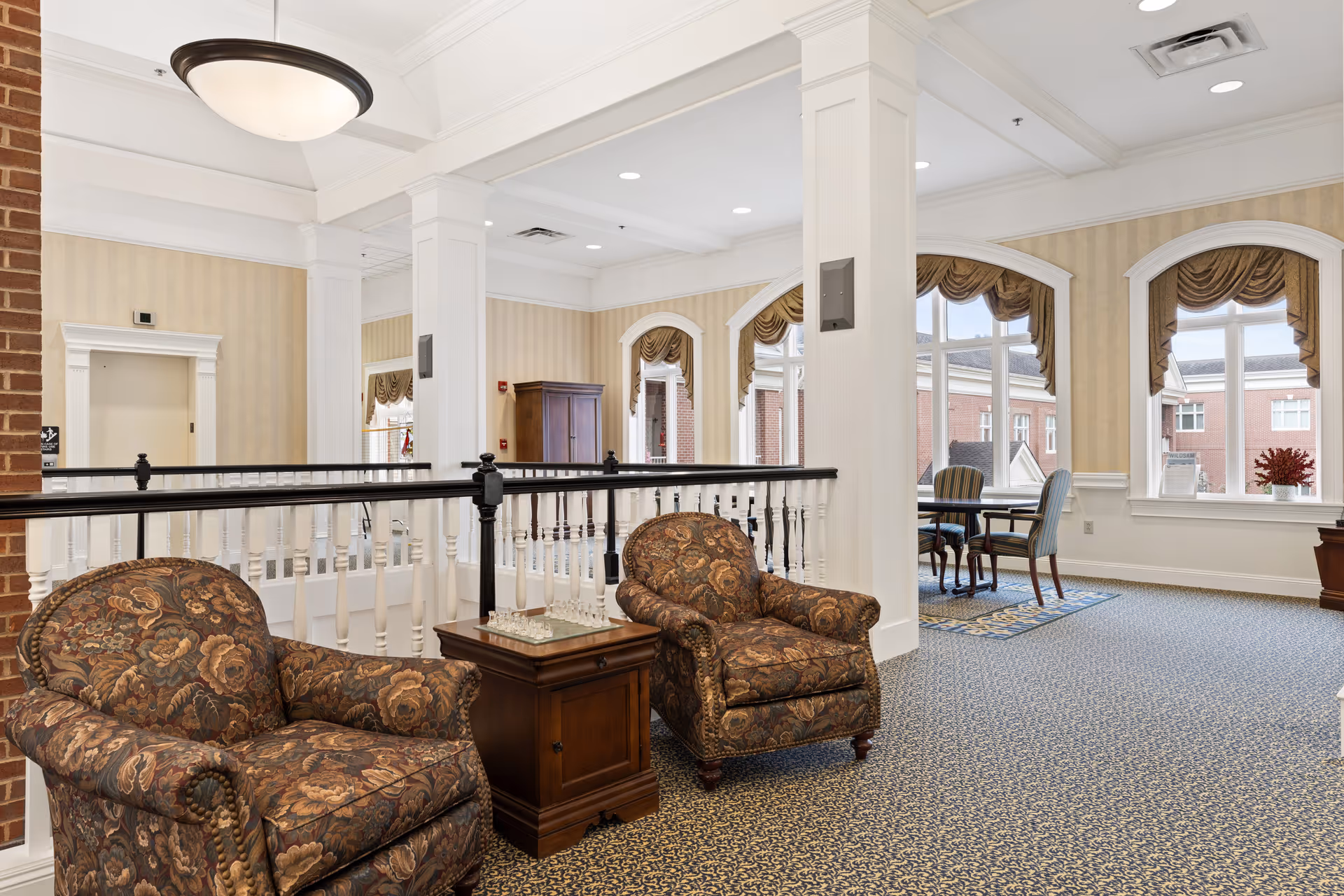 A bright and spacious sitting area in a senior living facility with two patterned armchairs and a wooden side table with a chess set on top. The room features large windows with decorative valances, beige striped wallpaper, white columns, and a patterned carpet. In the background, there is a small table with chairs near the windows.