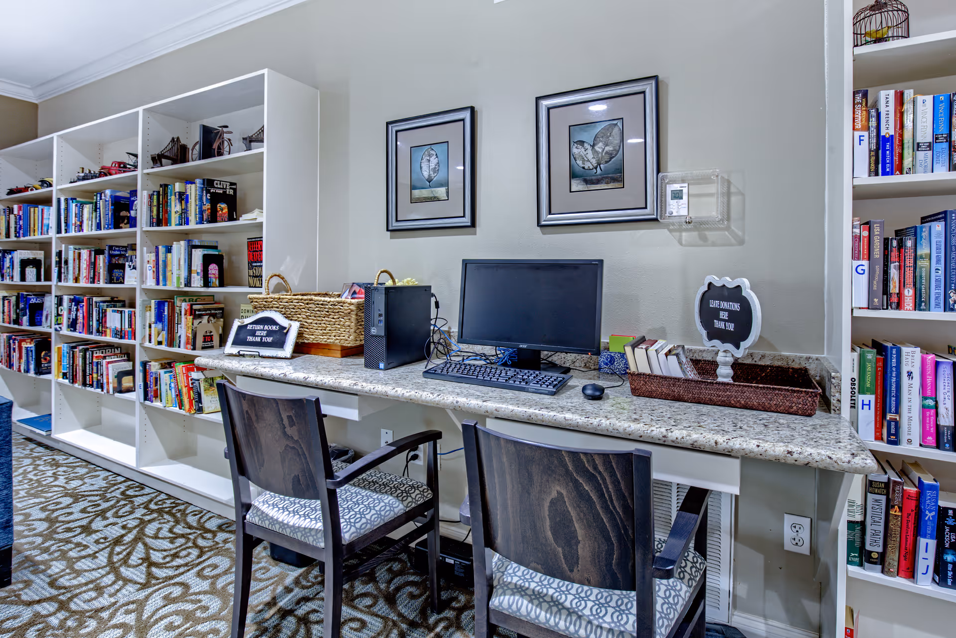 A cozy library or reading area with white bookshelves filled with books along the wall. In front of the shelves is a granite countertop desk with two wooden chairs. On the desk, there is a computer monitor, keyboard, mouse, a basket, and a small sign that says 'Return Books Here Thank You'. Two framed leaf artworks hang on the wall above the desk.