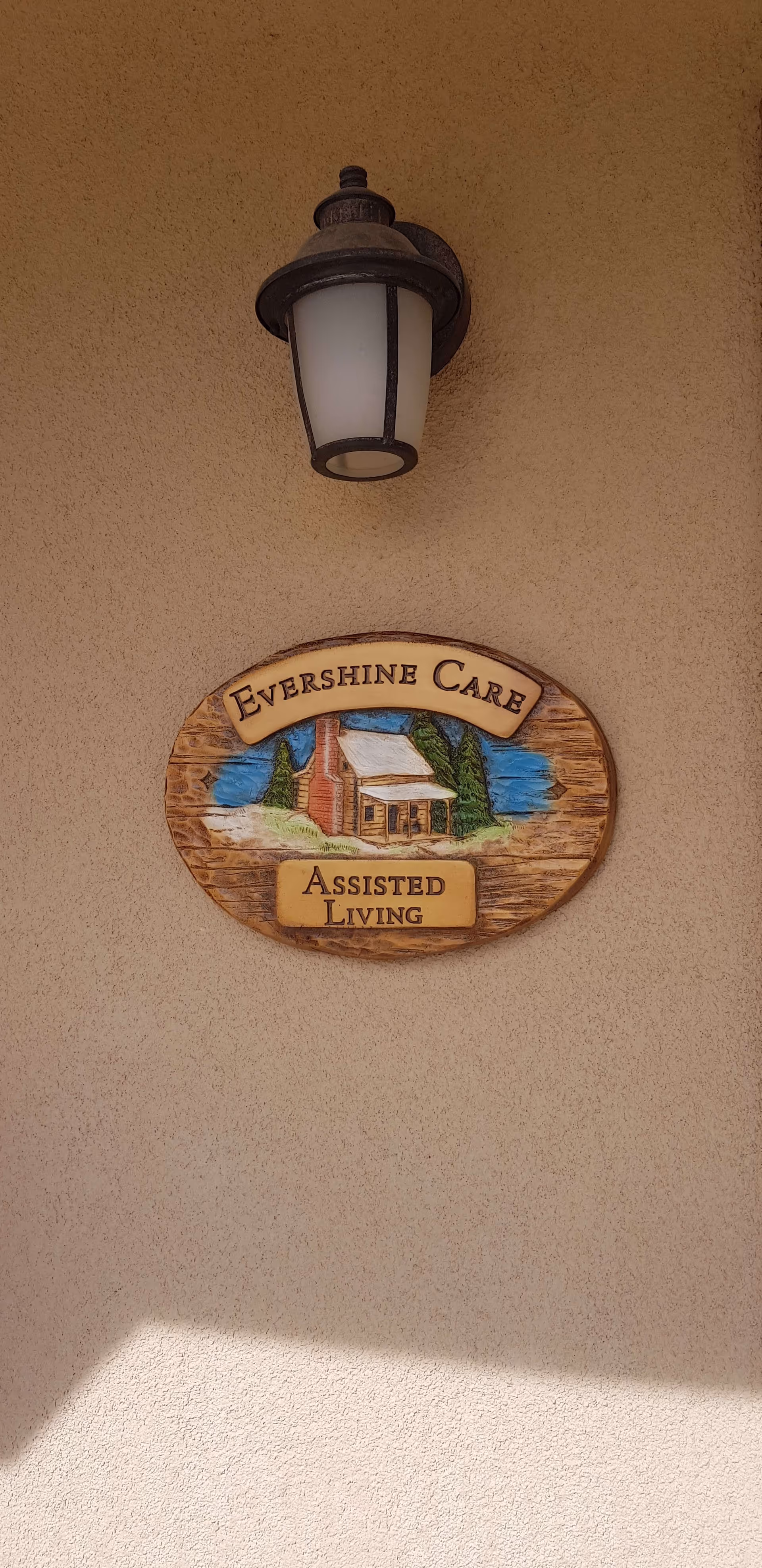 A wall-mounted light fixture above a decorative wooden sign that reads 'Evershine Care Assisted Living' with an illustration of a house and trees.
