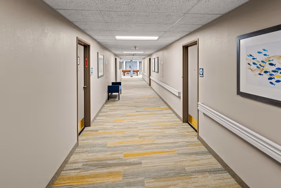 A long, well-lit hallway in a senior living facility with beige walls and a patterned carpet in shades of gray and yellow. There are several doors along both sides of the hallway, handrails on the walls, framed artwork, and a blue chair placed against the left wall. At the far end of the hallway, a pool table is visible.