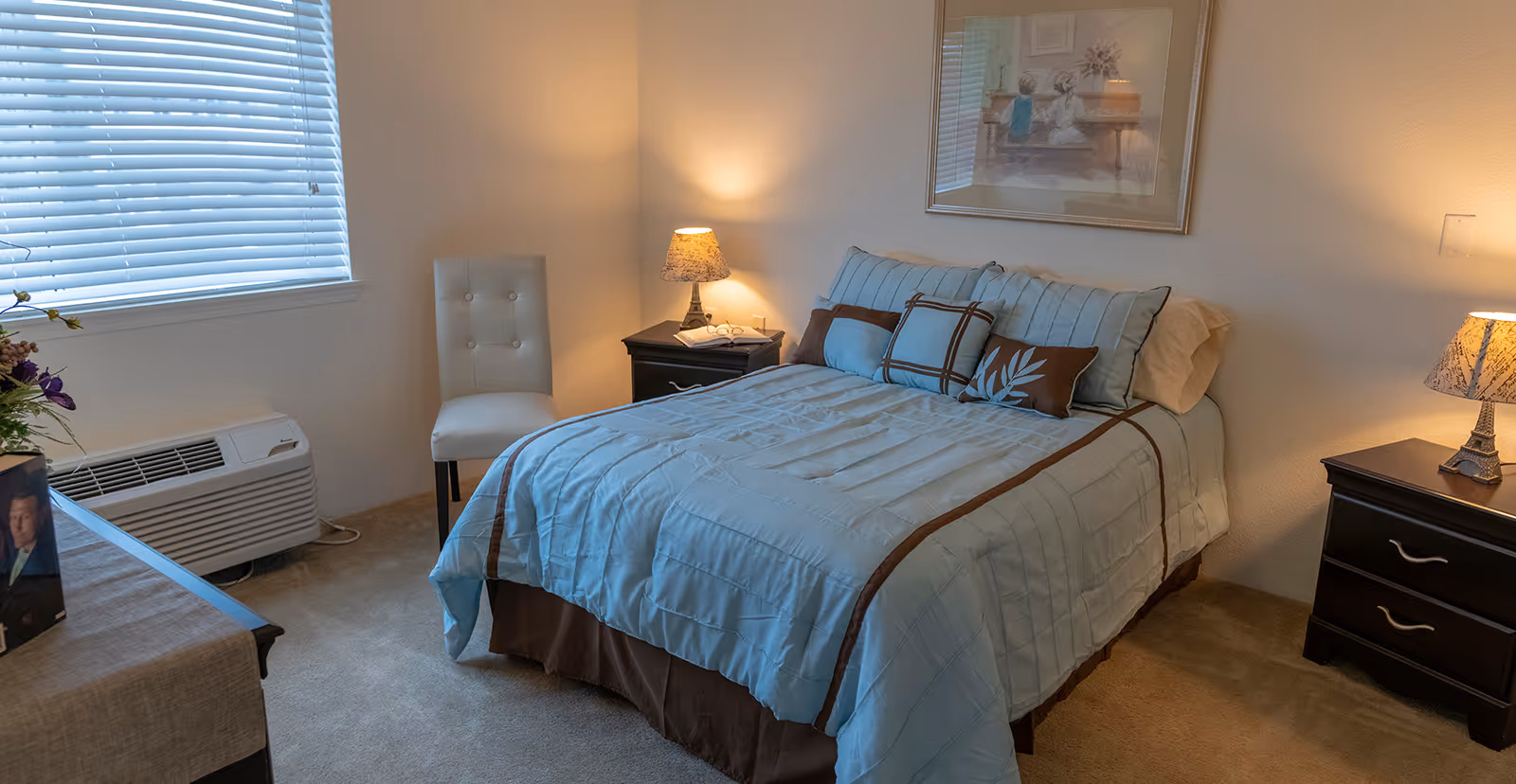 A cozy bedroom with a neatly made bed featuring light blue and brown bedding, two matching bedside tables with lamps, a white cushioned chair near a window with blinds, and a framed picture on the wall above the bed.