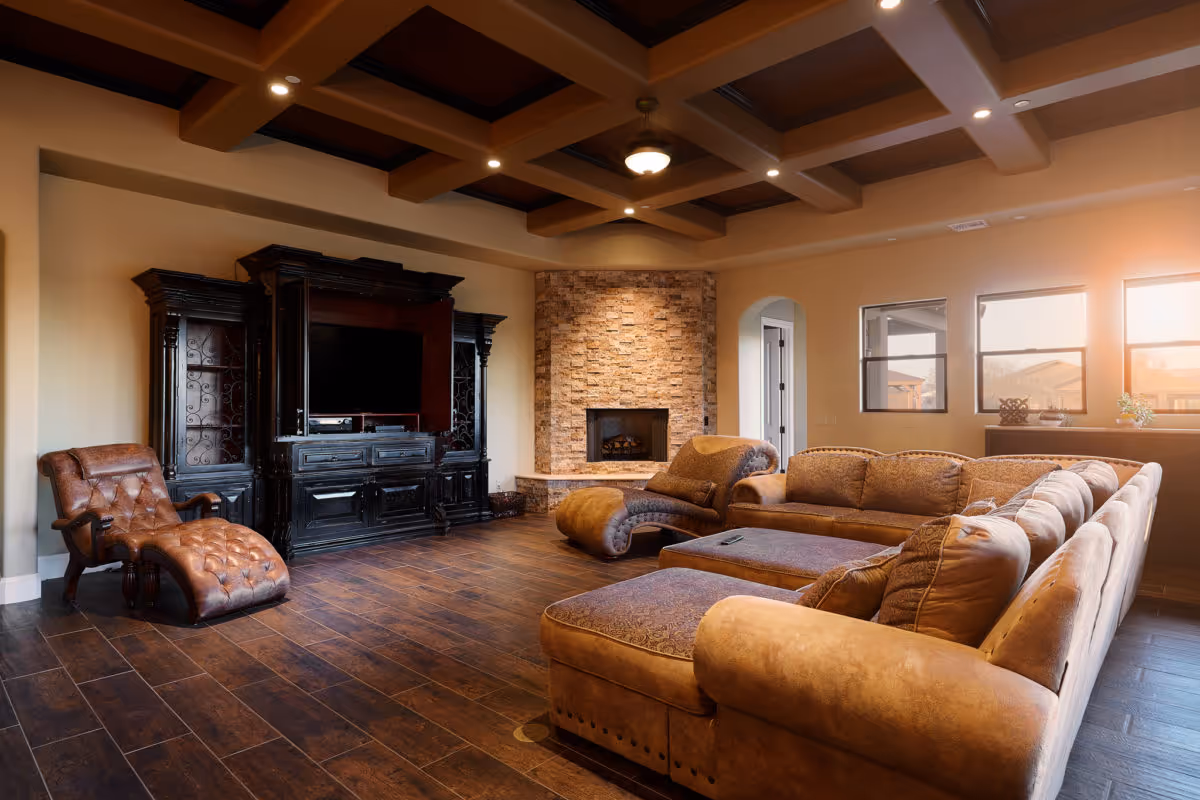 A spacious living room with a large brown sectional sofa, a matching chaise lounge chair, a dark wooden entertainment center with a TV, and a stone fireplace. The room has wooden flooring, exposed wooden beams on the ceiling, and three windows letting in natural light.
