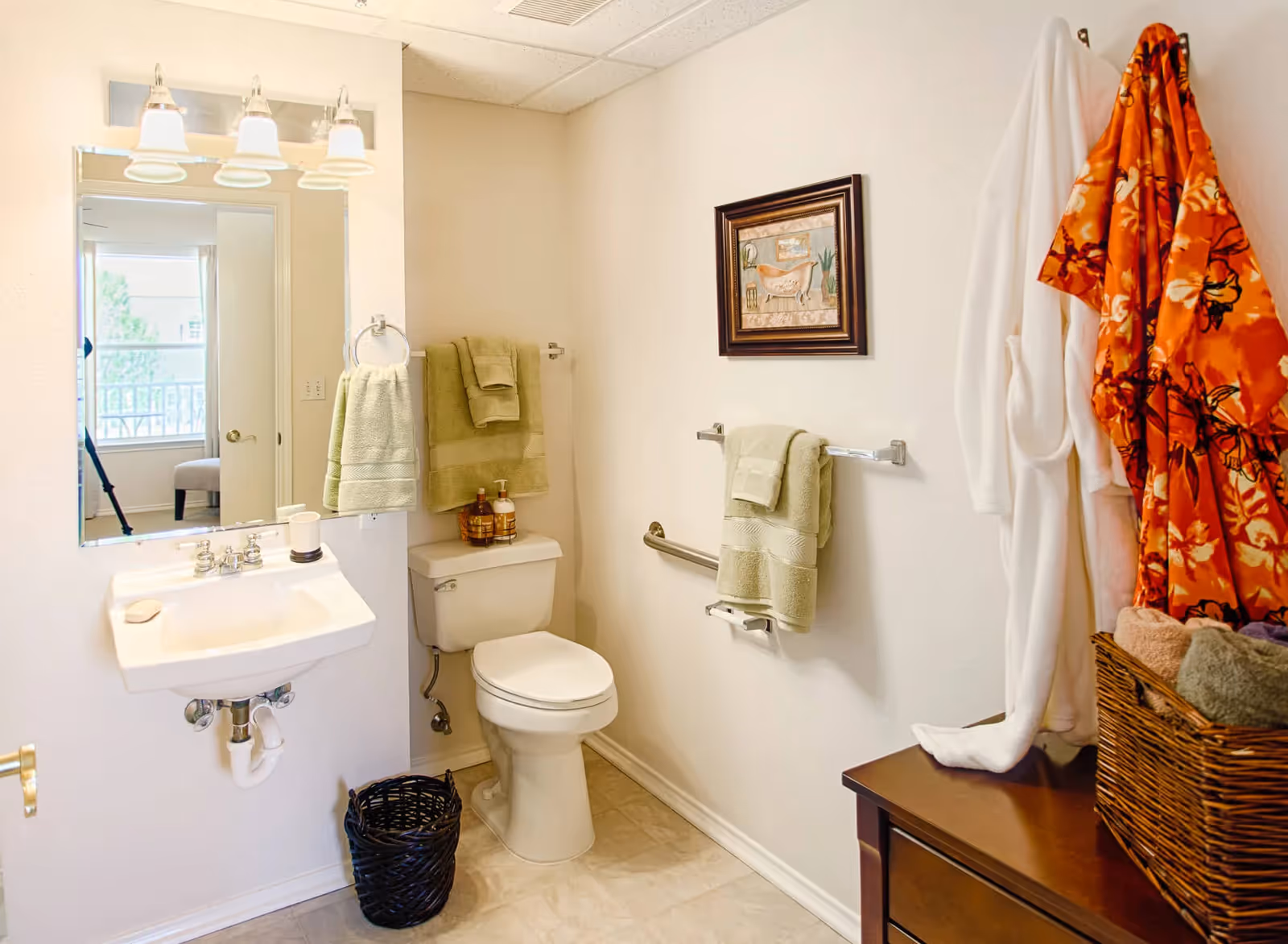 A clean and well-lit bathroom featuring a white toilet and a wall-mounted sink with a bar of soap. Above the sink is a large mirror with three light fixtures. Green towels hang on towel racks near the toilet, and a framed picture is mounted on the wall. To the right, a wooden cabinet holds a wicker basket with folded towels and two hanging robes, one white and one orange with a floral pattern. A black wastebasket is placed on the floor beneath the sink.