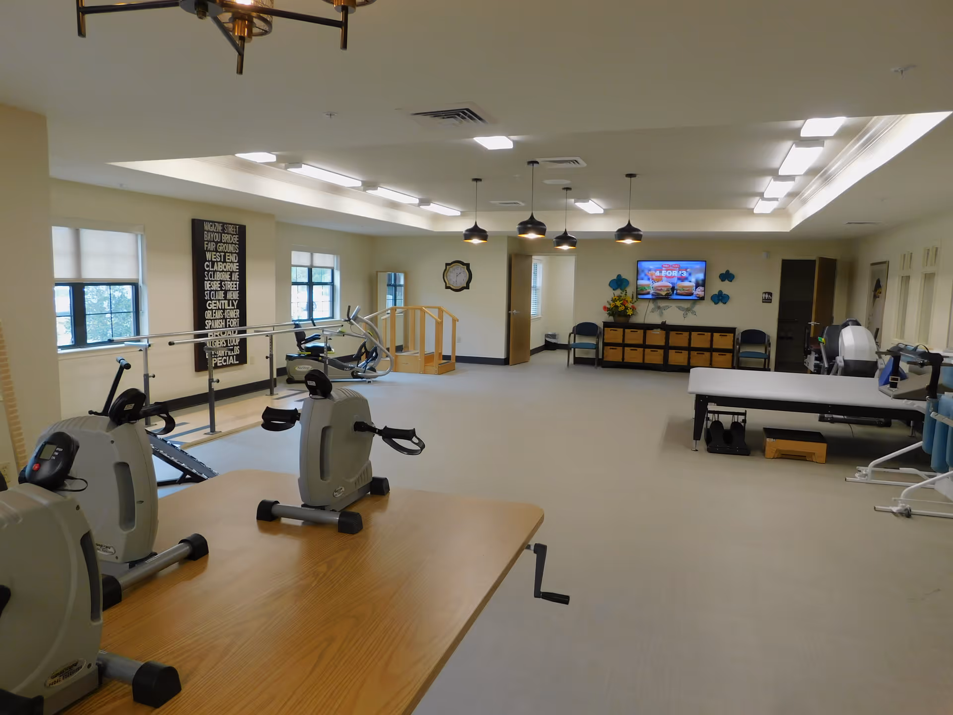 A spacious rehabilitation or physical therapy room with exercise equipment including stationary bikes, parallel bars for walking practice, a therapy table, and a TV mounted on the wall. The room has large windows, neutral-colored walls, and ceiling lights.