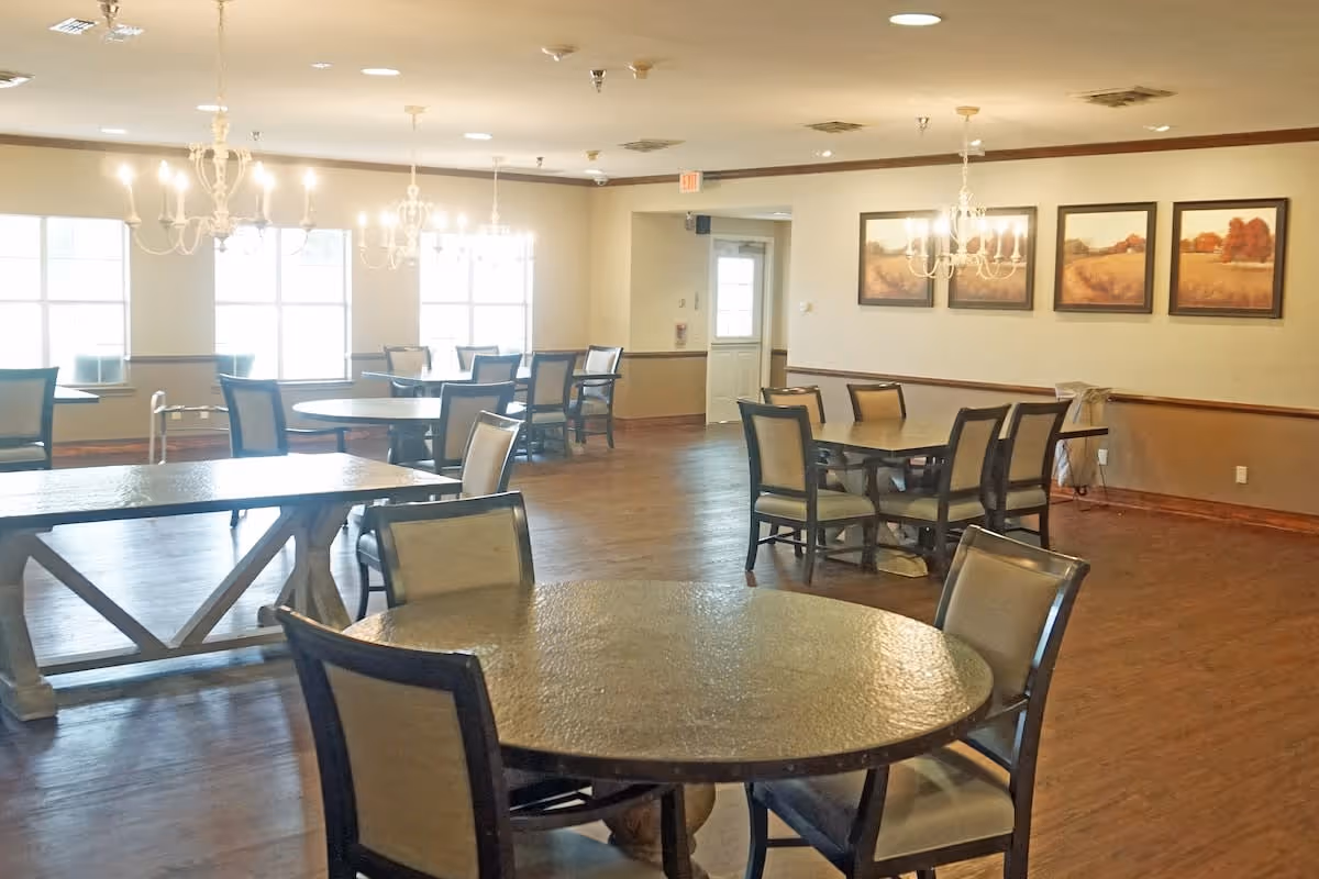 A spacious dining room with multiple round and rectangular tables surrounded by chairs. The room has wooden flooring, several chandeliers hanging from the ceiling, large windows letting in natural light, and framed landscape paintings on the wall.