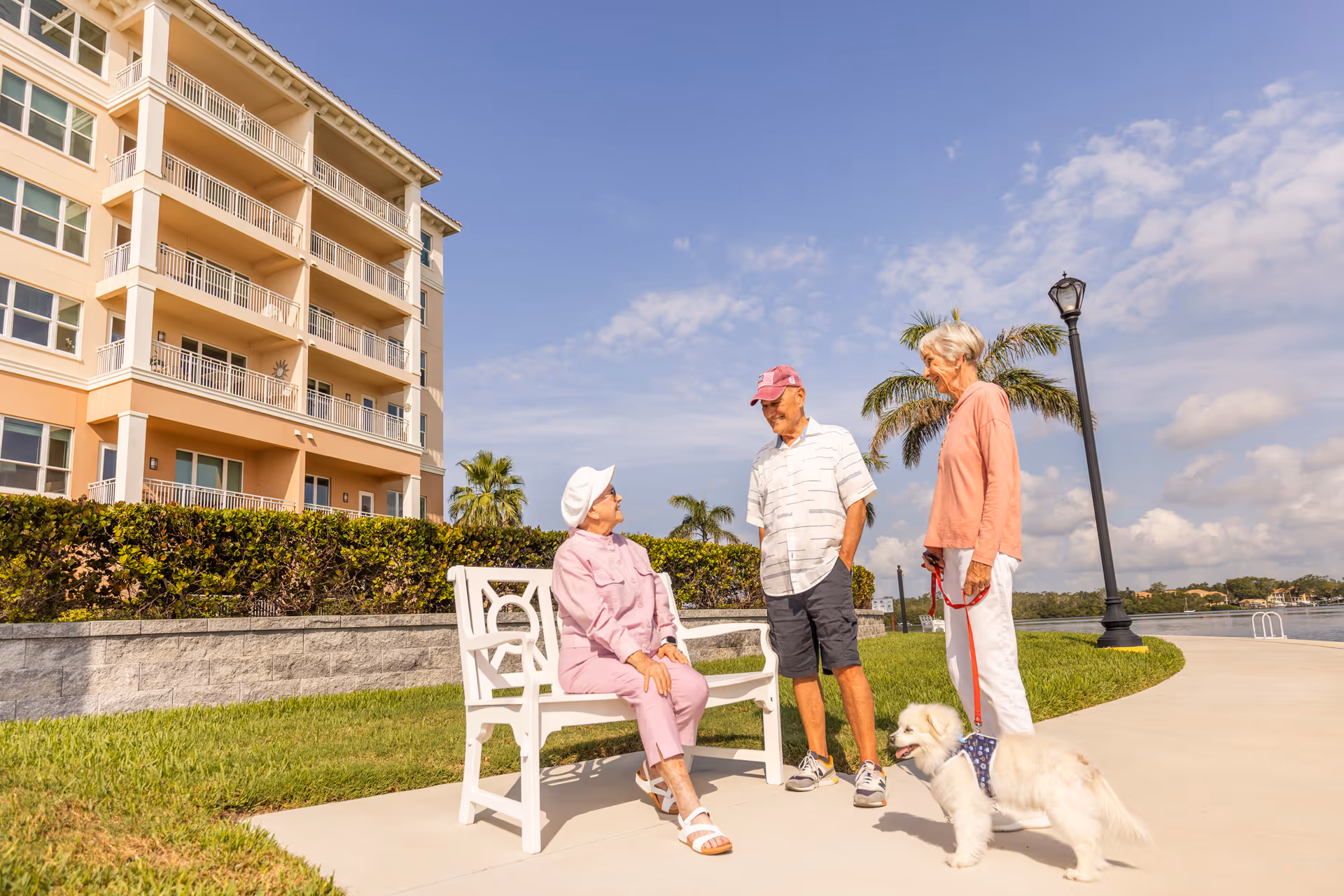 Three elderly people outdoors near a waterfront with a multi-story residential building in the background. One woman is sitting on a white bench wearing a pink outfit and white hat, while a man in a white shirt and red cap and another woman in a peach top holding a small white dog on a leash stand nearby. Palm trees and a lamppost are visible under a partly cloudy sky.