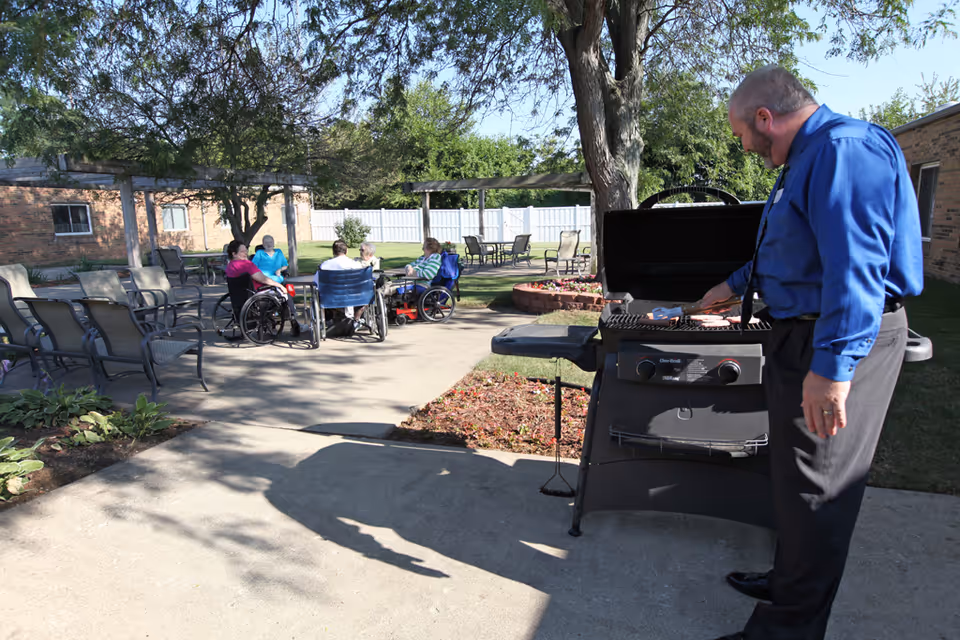 A man in a blue shirt and black pants is grilling food on a barbecue grill outdoors. In the background, a group of elderly people, some in wheelchairs, are sitting and chatting around a table in a garden area with chairs and trees. The setting appears to be a sunny day at a senior living facility.
