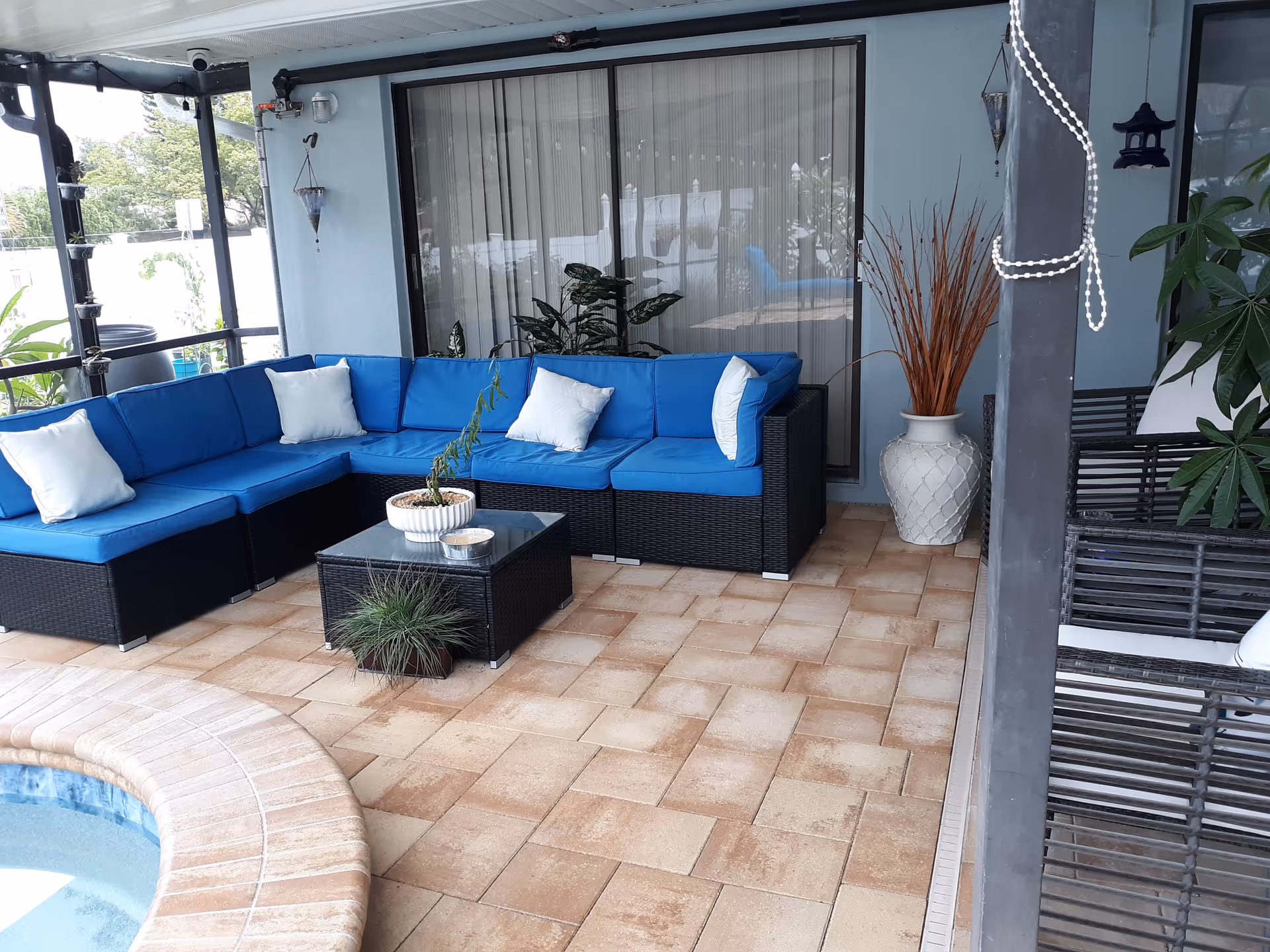 Covered outdoor patio area with a blue cushioned sectional sofa, white pillows, a glass-top coffee table with potted plants, and a small pool edge visible in the foreground. The patio has tiled flooring and is decorated with hanging planters and a large vase with dried branches.