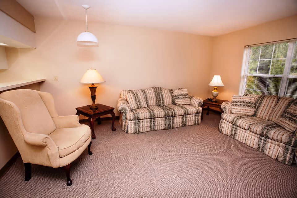 A cozy living room with two patterned sofas and a beige armchair arranged around two wooden side tables, each with a lamp. The room has beige walls, a carpeted floor, a window with blinds, and a hanging ceiling light.