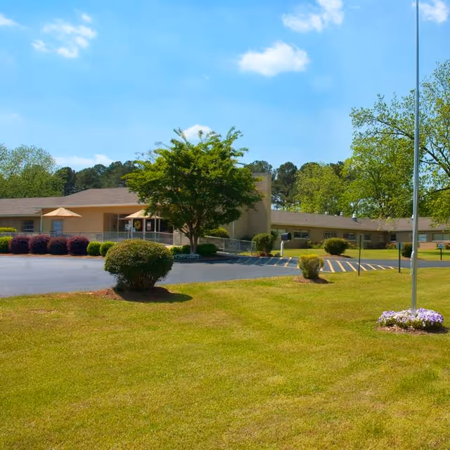 Single-story senior living facility building with a driveway, landscaped lawn, trees, and a flagpole under a blue sky.