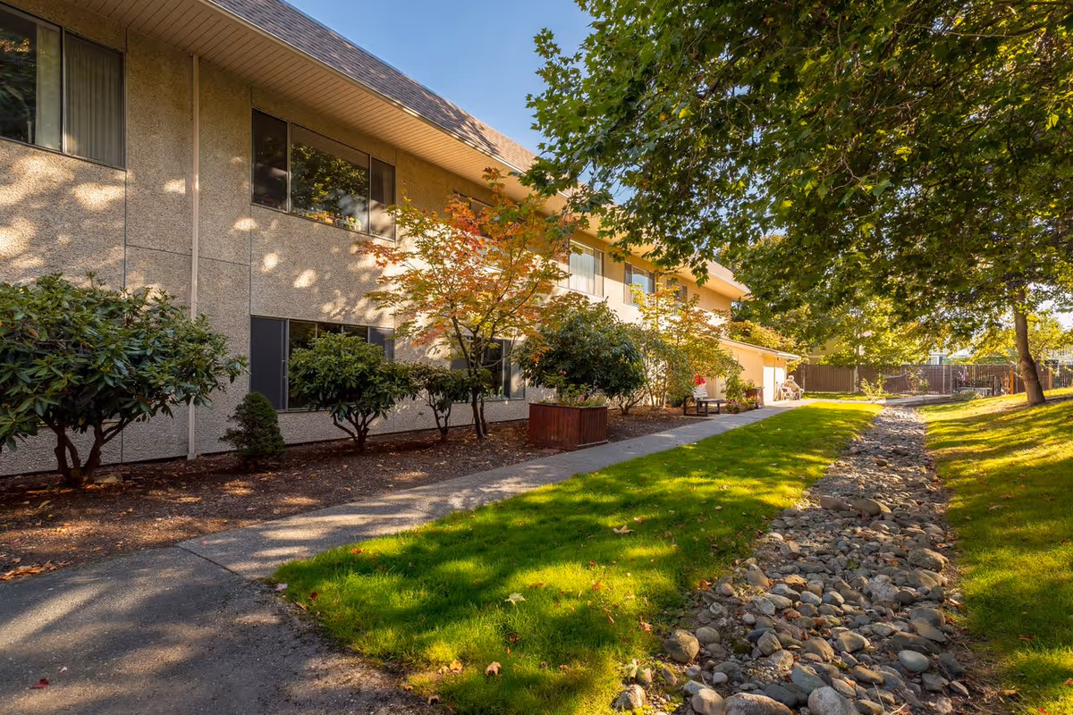 Exterior view of Rosewood Villa's landscaped grounds with a walkway, grassy lawn, rock drainage bed, and a two-story building shaded by trees.
