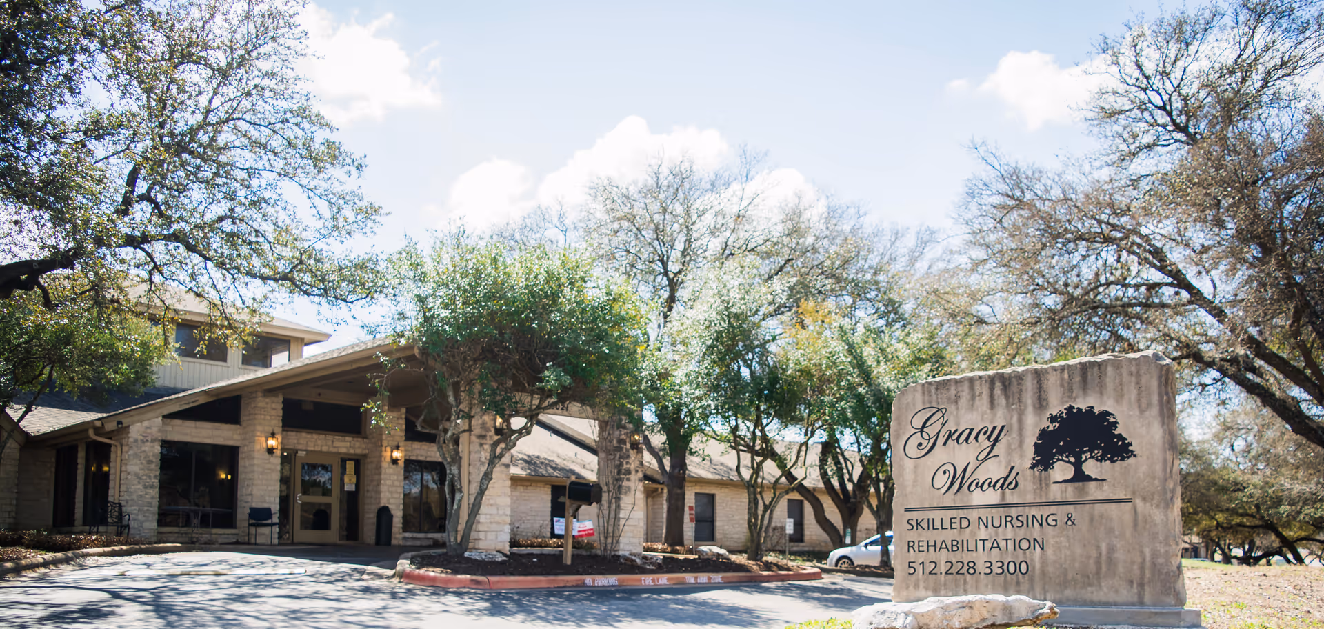 Exterior view of Gracy Woods I Nursing Center showing the entrance of the building with stone walls and large windows, surrounded by trees under a partly cloudy sky. A stone sign in the foreground displays the facility's name, services, and phone number.
