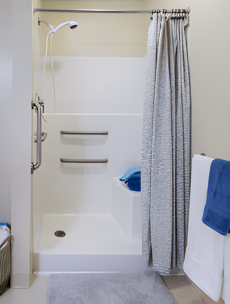 A clean, accessible shower area with a white shower base, two built-in shelves, a handheld showerhead, and a gray shower curtain partially drawn to the side. A blue towel is folded on a built-in seat inside the shower, and a white towel with a blue towel on top hangs on a rack outside the shower. A gray bath mat is placed on the floor in front of the shower.