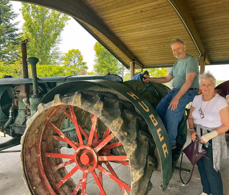 An elderly man sitting on a vintage green and red Huber tractor under a wooden shelter, with an elderly woman standing beside him holding a purse and wearing a wrist brace. Trees and greenery are visible in the background.