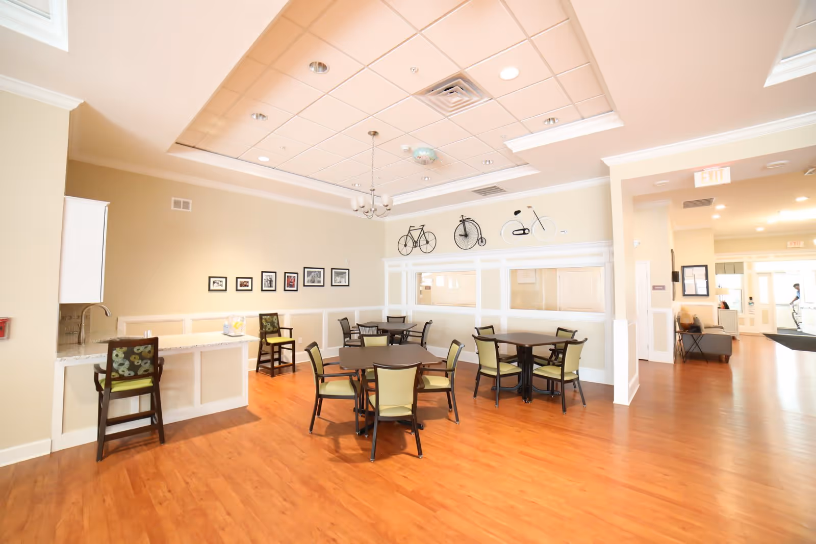 A bright and spacious common area with wooden flooring, several tables with chairs arranged around them, a small counter with two chairs, framed pictures on the wall, and decorative bicycle wall art above a white panel wall. The area has a high ceiling with recessed lighting and a chandelier, and an exit sign is visible in the background.