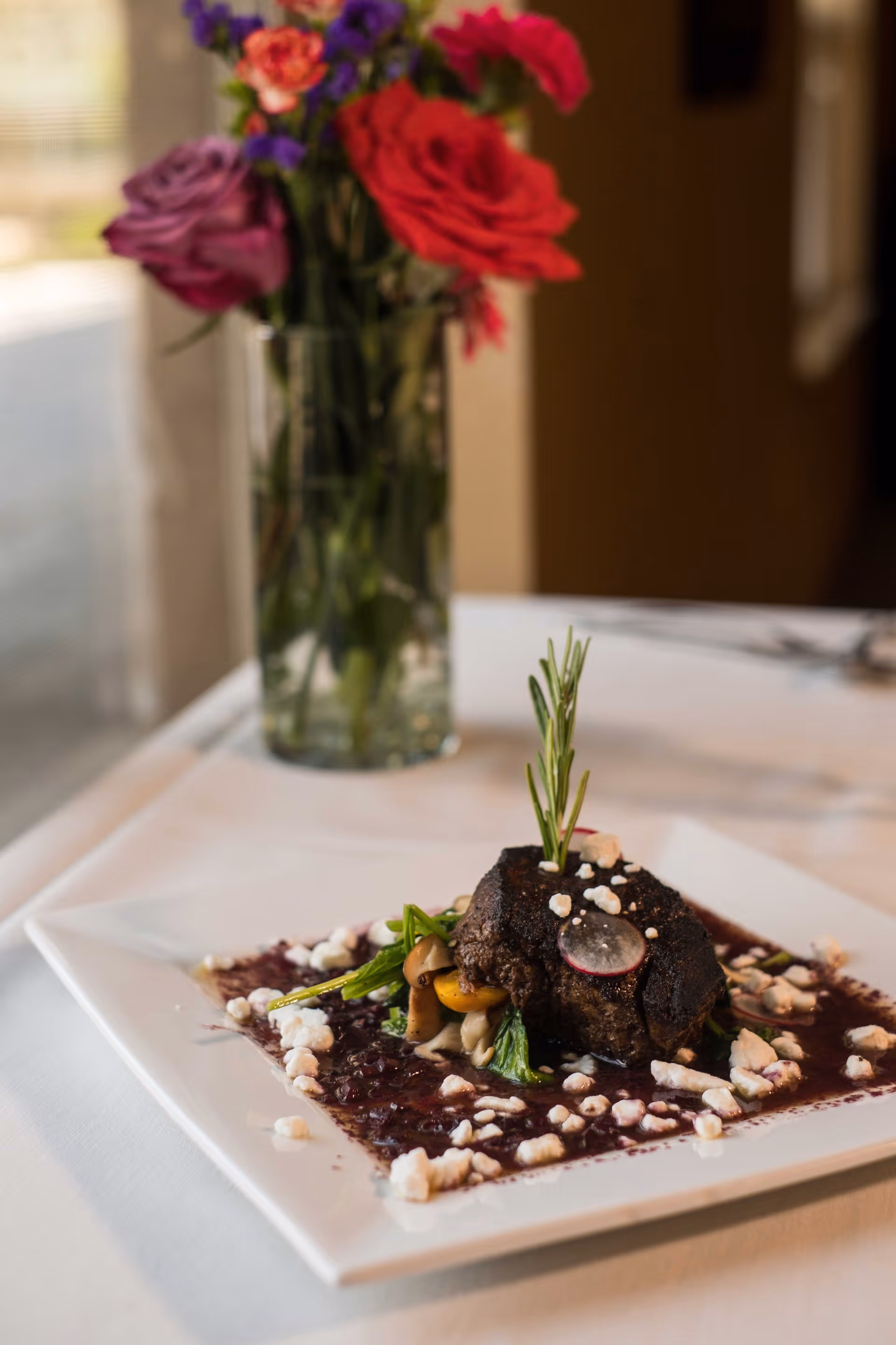 A gourmet plated dish featuring a piece of cooked meat garnished with a sprig of rosemary, radish slice, mushrooms, and greens, served on a white square plate with a dark sauce and white crumbles. In the background, there is a vase with colorful flowers on a white tablecloth.