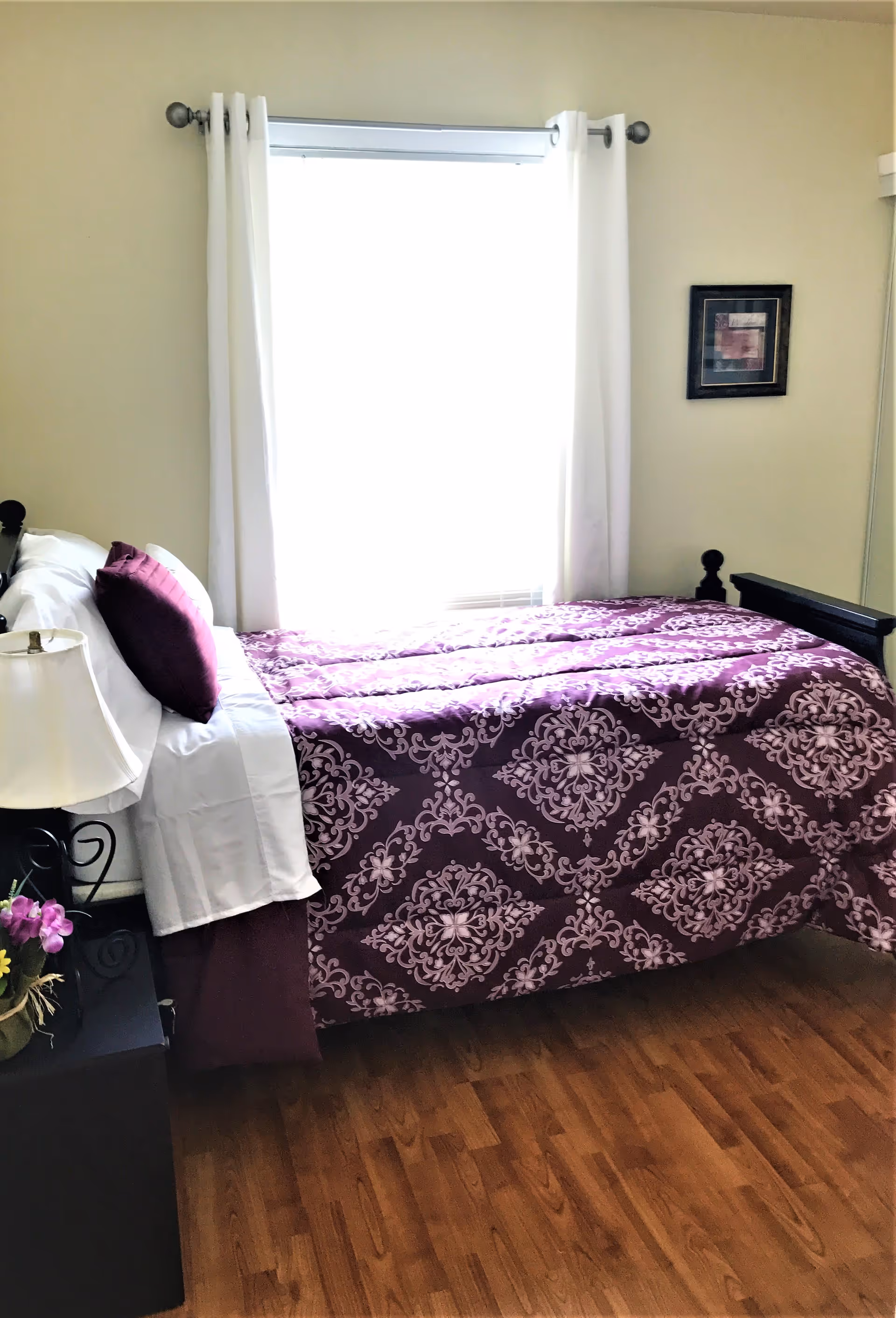A bedroom with a bed covered in a purple and white patterned comforter and matching pillows. There is a window with white curtains behind the bed, a bedside table with a lamp and a small flower arrangement, and a framed picture on the wall.