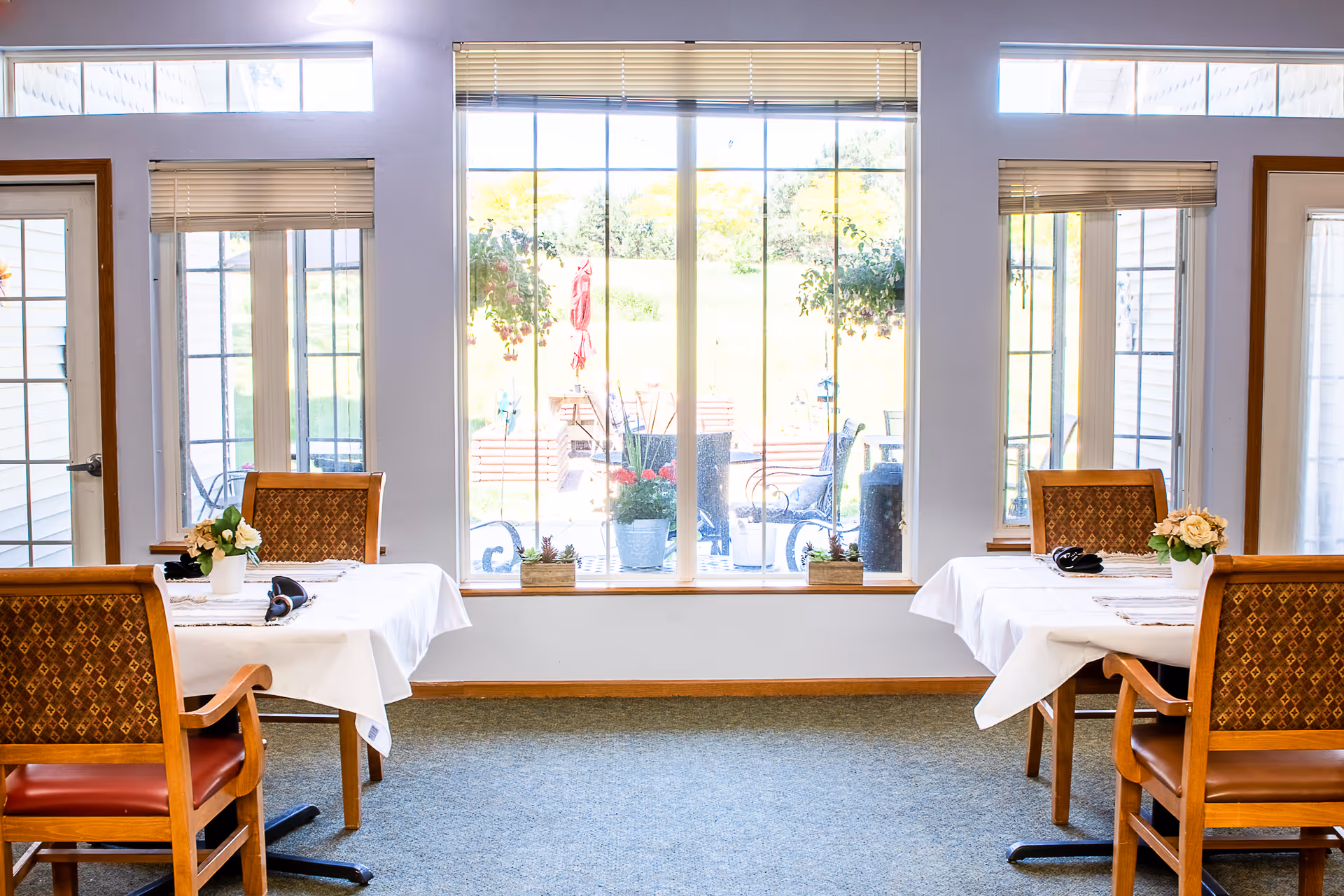 A bright dining room with two tables covered in white tablecloths, each set with a small flower arrangement and headphones. Four wooden chairs with patterned upholstery surround the tables. Large windows and glass doors let in natural light and offer a view of an outdoor patio with chairs and plants.