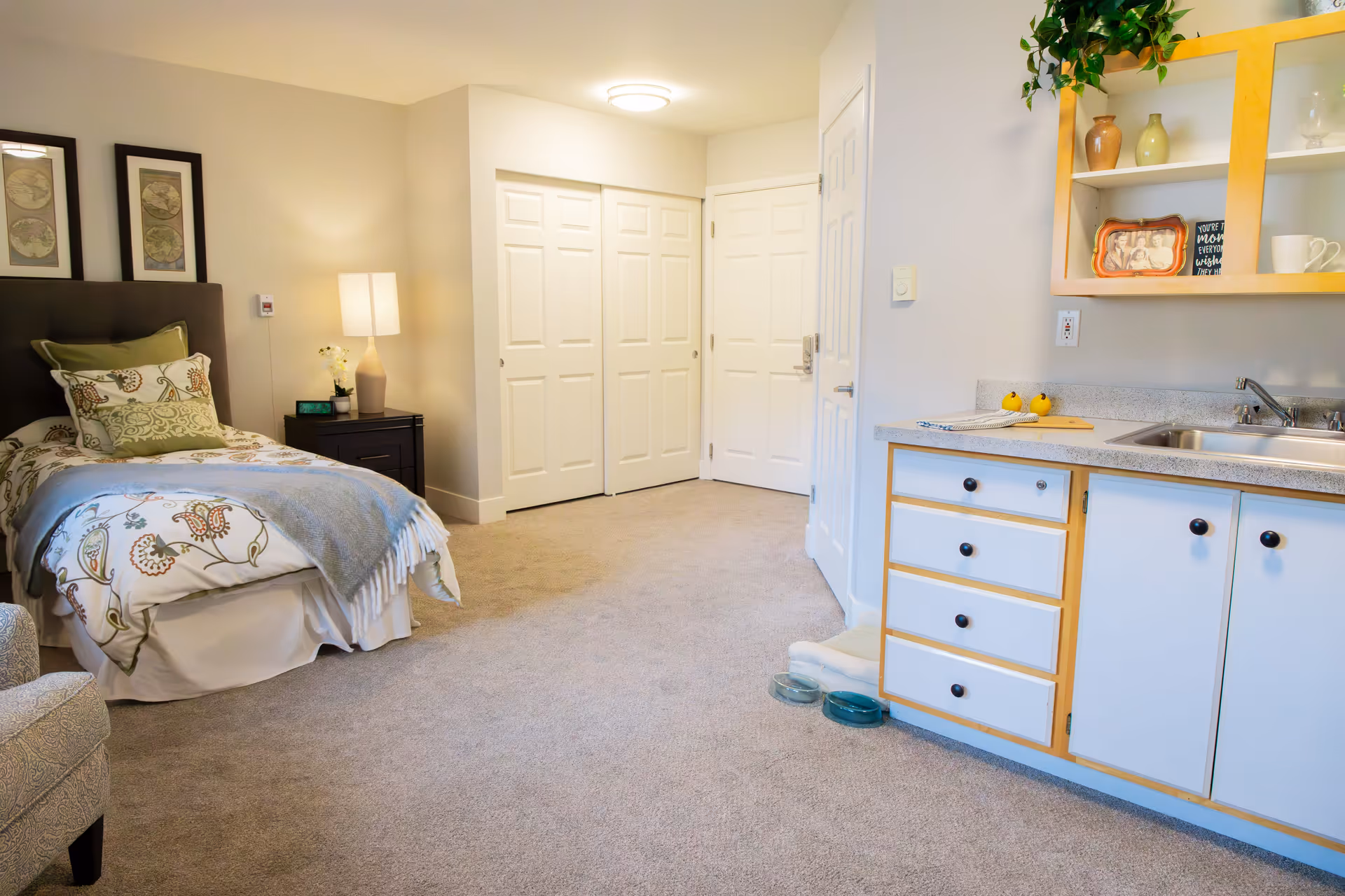 A bright and clean senior living studio apartment featuring a single bed with patterned bedding and multiple pillows, a nightstand with a lamp and a small plant, a kitchenette with a sink, cabinets, and countertop, and a closet with white double doors. The room has beige carpet and neutral-colored walls, creating a cozy and welcoming atmosphere.
