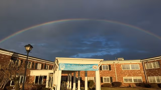 Exterior view of a two-story brick building with a covered entrance and a blue banner that reads 'St Patrick's Manor'. A street lamp is visible on the left side, and a rainbow arches across the cloudy sky above the building.