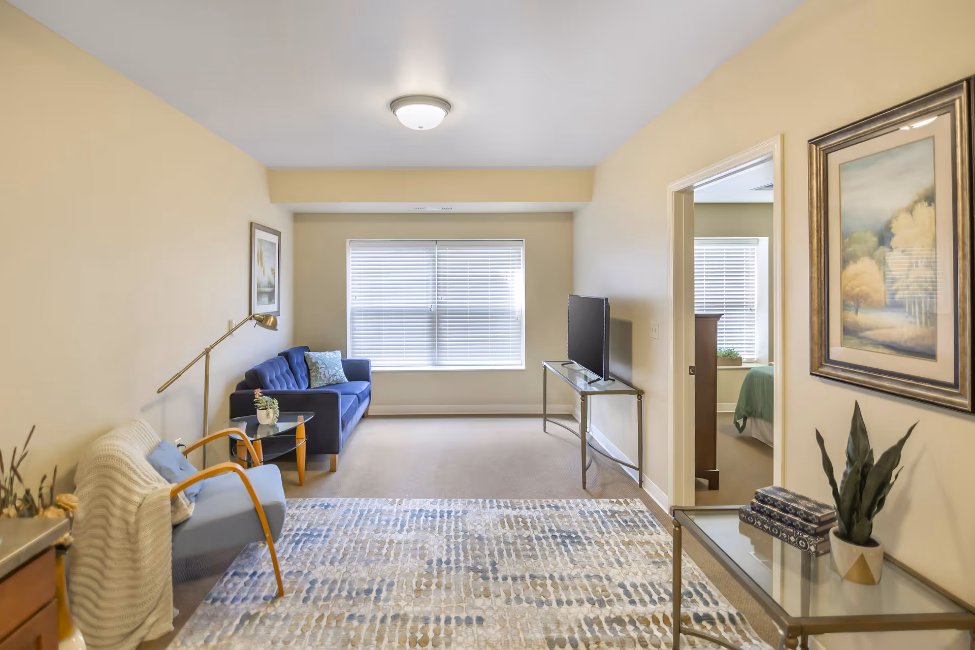 A cozy living room with beige walls and carpet, featuring a blue sofa with a decorative pillow, a light blue armchair with a white knitted throw, a glass side table with a small plant, a floor lamp, and a flat-screen TV on a glass stand. A doorway leads to a bedroom with a green bedspread, and a framed landscape painting hangs on the wall near the doorway.