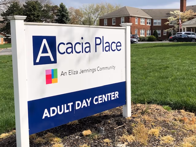 Outdoor view of a sign for Acacia Place Adult Day Center, an Eliza Jennings Community, with a large brick building and green lawn in the background under a partly cloudy sky.