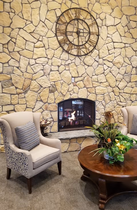 Cozy seating area with two upholstered armchairs featuring patterned sides and striped cushions, a round wooden coffee table with a floral arrangement, and a stone wall with a fireplace and a large decorative clock above it.