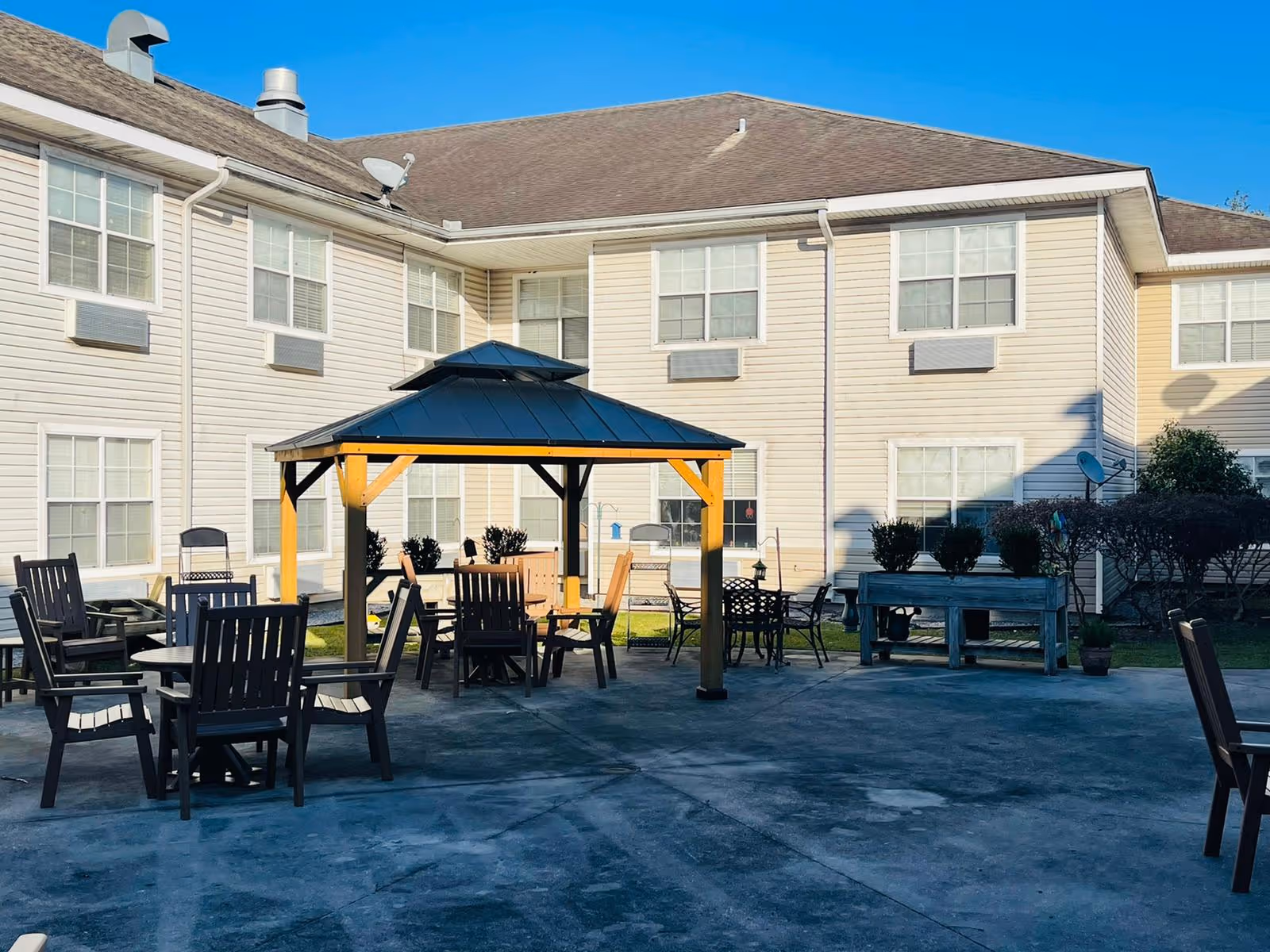 Outdoor courtyard area of a senior living facility with a gazebo featuring a black roof and wooden supports. Several wooden chairs and tables are arranged around the gazebo on a concrete patio. The background shows a two-story beige building with multiple windows and some shrubs along the building's edge under a clear blue sky.