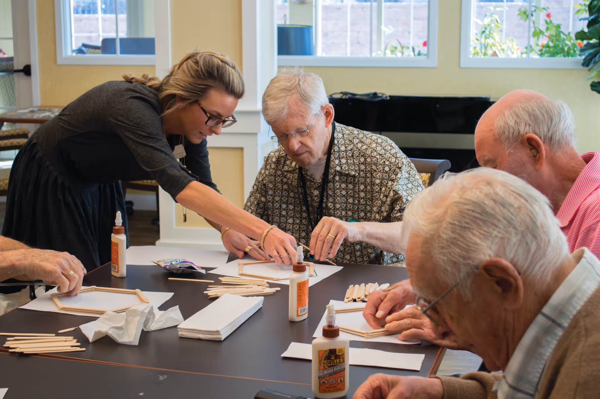 A group of elderly men sitting around a table working on a craft project with wooden sticks and glue, assisted by a younger woman in a bright room with large windows.