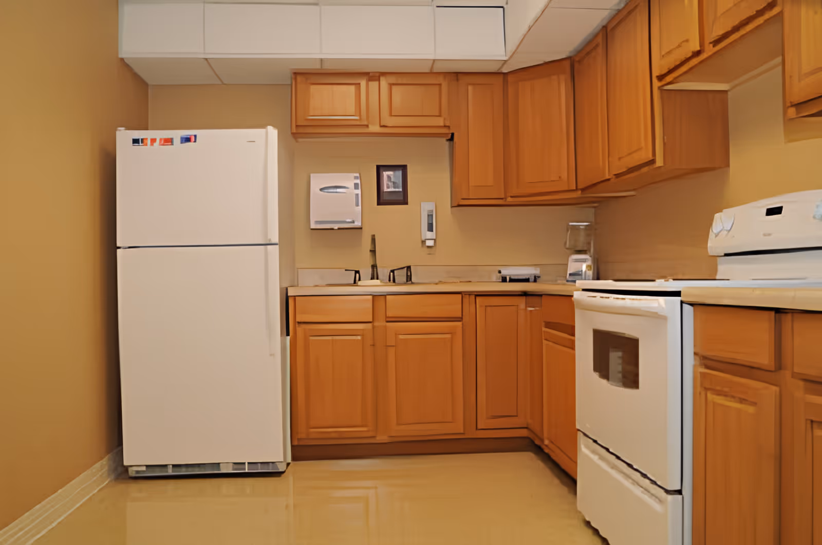 A kitchen with wooden cabinets, a white refrigerator, a white stove, a blender on the countertop, a sink with two faucets, and a wall-mounted paper towel dispenser and soap dispenser.