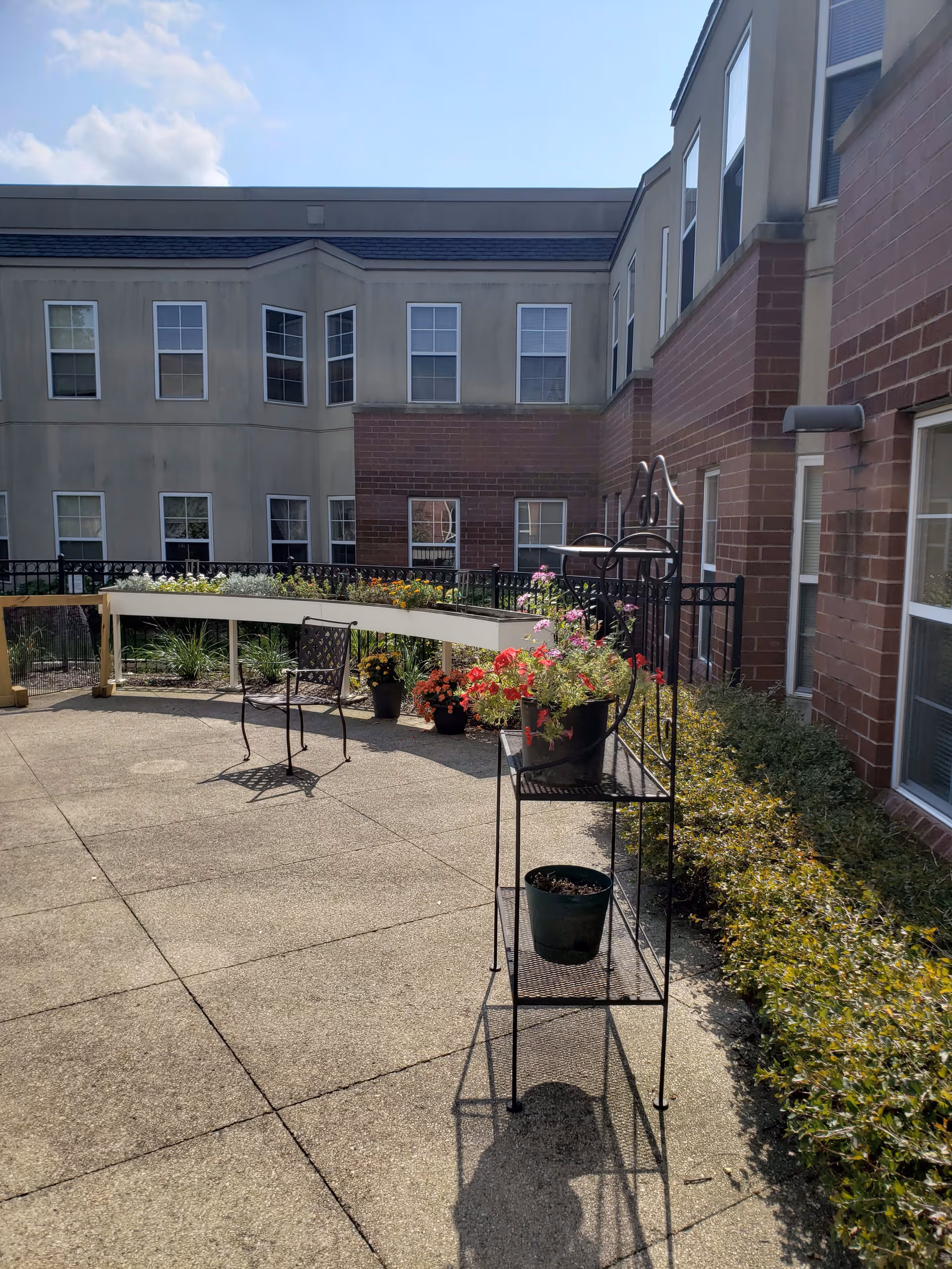 Outdoor courtyard area of a senior living facility with a concrete patio, metal plant stand holding flower pots, a metal chair, and various potted plants and flowers along the perimeter. The courtyard is surrounded by a two-story building with multiple windows and brick and beige exterior walls under a partly cloudy sky.