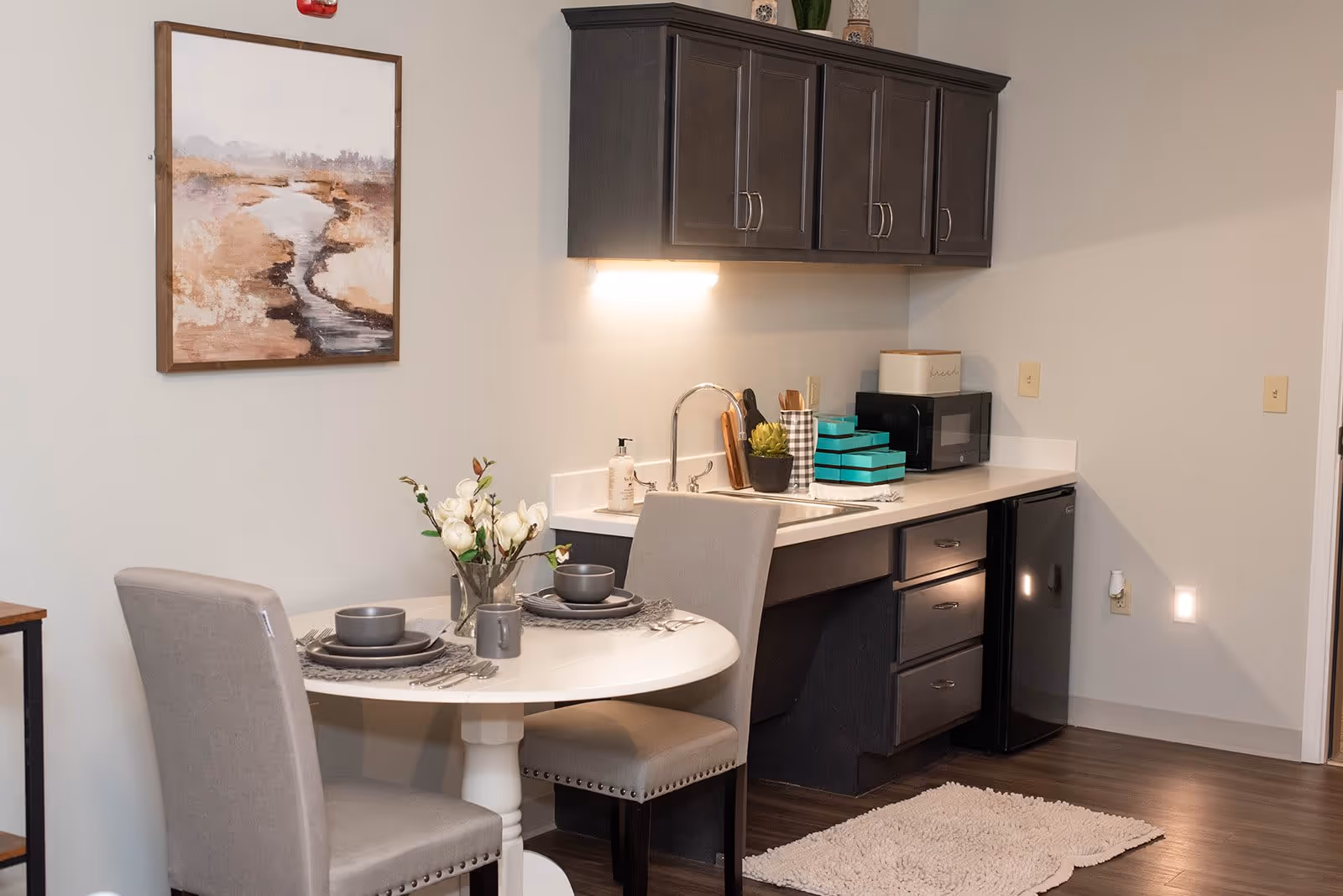 A small kitchenette area with dark upper and lower cabinets, a sink, a microwave, and a mini refrigerator. In front of the kitchenette is a round white table set for two with gray chairs, gray dishware, and a vase with white flowers. A framed landscape painting hangs on the wall above the table.