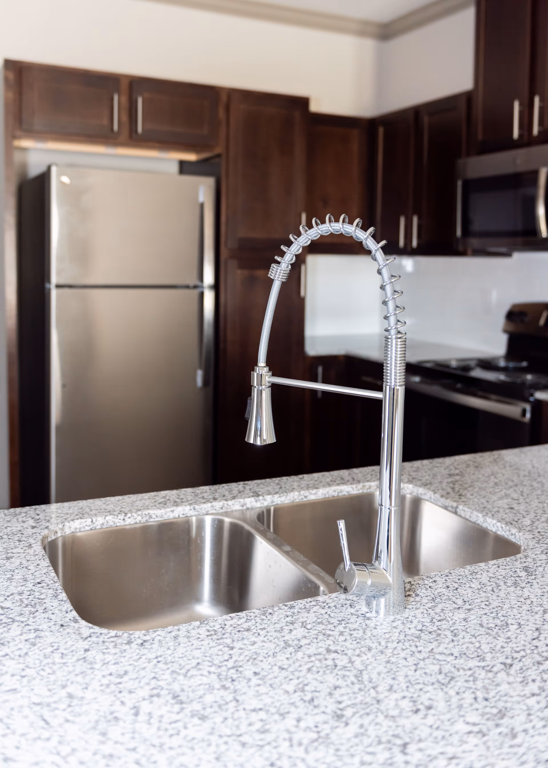 Modern kitchen interior featuring a double stainless steel sink with a high-arc spring coil faucet on a speckled granite countertop. In the background, there are dark wooden cabinets, a stainless steel refrigerator, a microwave, and a stove.