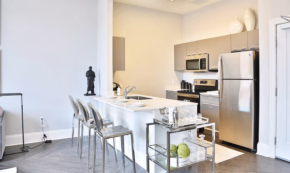 Modern kitchen area with stainless steel refrigerator, microwave, and stove. A white countertop island with a sink and three transparent bar stools. A small metal cart with glassware and decorative green artichokes is placed next to the island. Light-colored walls and flooring with minimalistic decor.