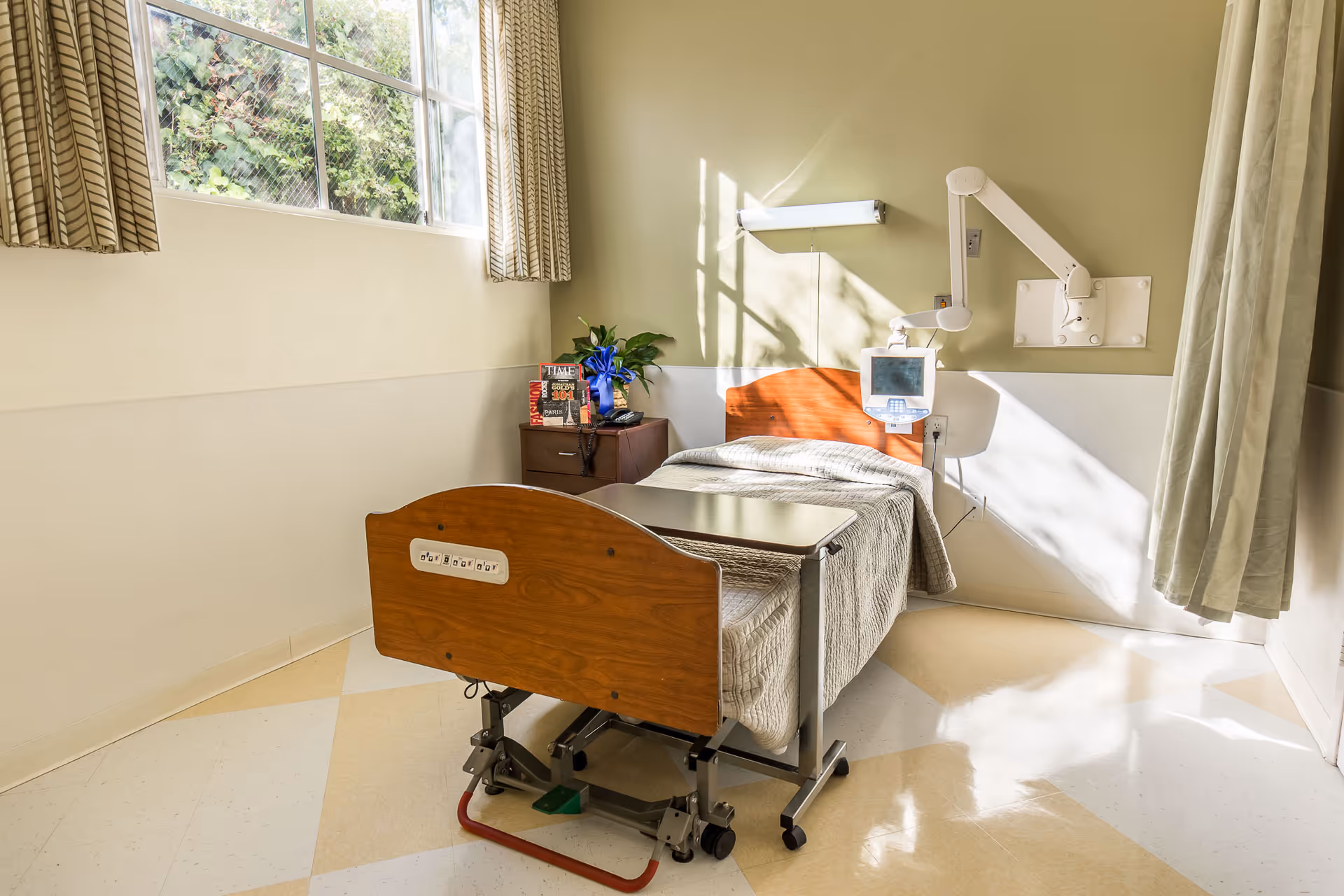 A small, sunlit hospital room with a single adjustable bed covered with a beige blanket. Next to the bed is a bedside table with a plant, a telephone, and some magazines. A medical monitor is mounted on the wall above the bed. The room has a large window with patterned curtains and light-colored walls and floor.