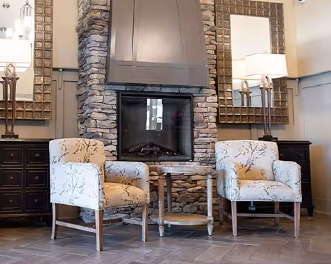 A cozy seating area with two patterned armchairs facing a stone fireplace. Between the chairs is a small wooden table. On either side of the fireplace are dark wooden cabinets topped with tall lamps and large decorative mirrors hanging above them.