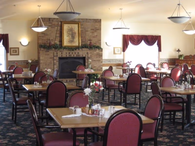 A dining room with multiple tables and chairs arranged neatly. Each table has a small flower vase and place settings. The room features a brick fireplace with a framed picture above it, wall sconces, hanging light fixtures, and windows with burgundy curtains.