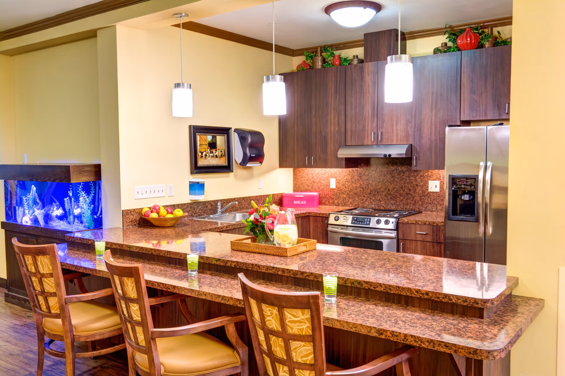 A modern kitchen area with a granite countertop bar and three wooden chairs with cushioned seats. The kitchen features dark wood cabinets, a stainless steel refrigerator, oven, and stove. Above the counter are three pendant lights. There is a bowl of apples, a pink bread box, a framed picture on the wall, and a fish tank with blue lighting on the left side. A tray with a pitcher of lemonade and flowers is placed on the counter.
