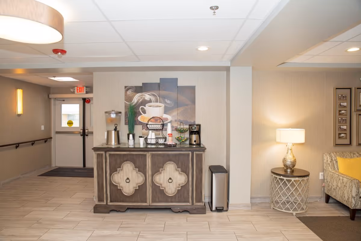 Interior view of a common area in an assisted living facility featuring a wooden cabinet with a coffee station including a coffee maker, water dispenser, cups, and condiments. Above the cabinet is a multi-panel wall art depicting a steaming cup of coffee. To the right, there is a round side table with a decorative lamp and part of a patterned armchair with a yellow cushion. The area has tiled flooring, beige walls, and ceiling lights. An exit door is visible in the background.