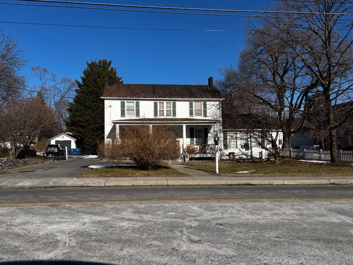 Two-story white house with a front porch, driveway, leafless trees, and a street in the foreground under a clear blue sky.