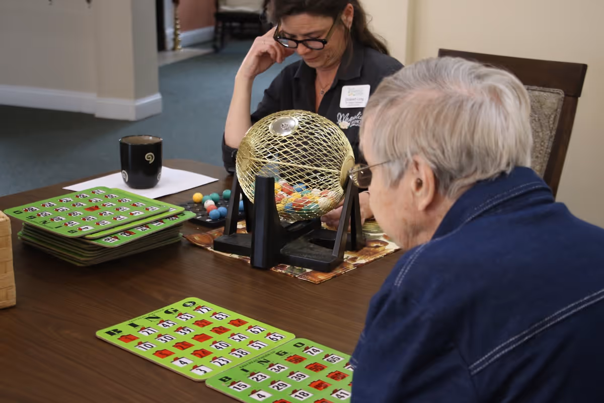 An elderly person and a staff member at Magnolia Manor of St. Marys are playing bingo at a wooden table. The elderly person is focused on the bingo cards in front of them, while the staff member is operating the bingo cage with numbered balls. A coffee mug and a stack of bingo cards are also on the table.