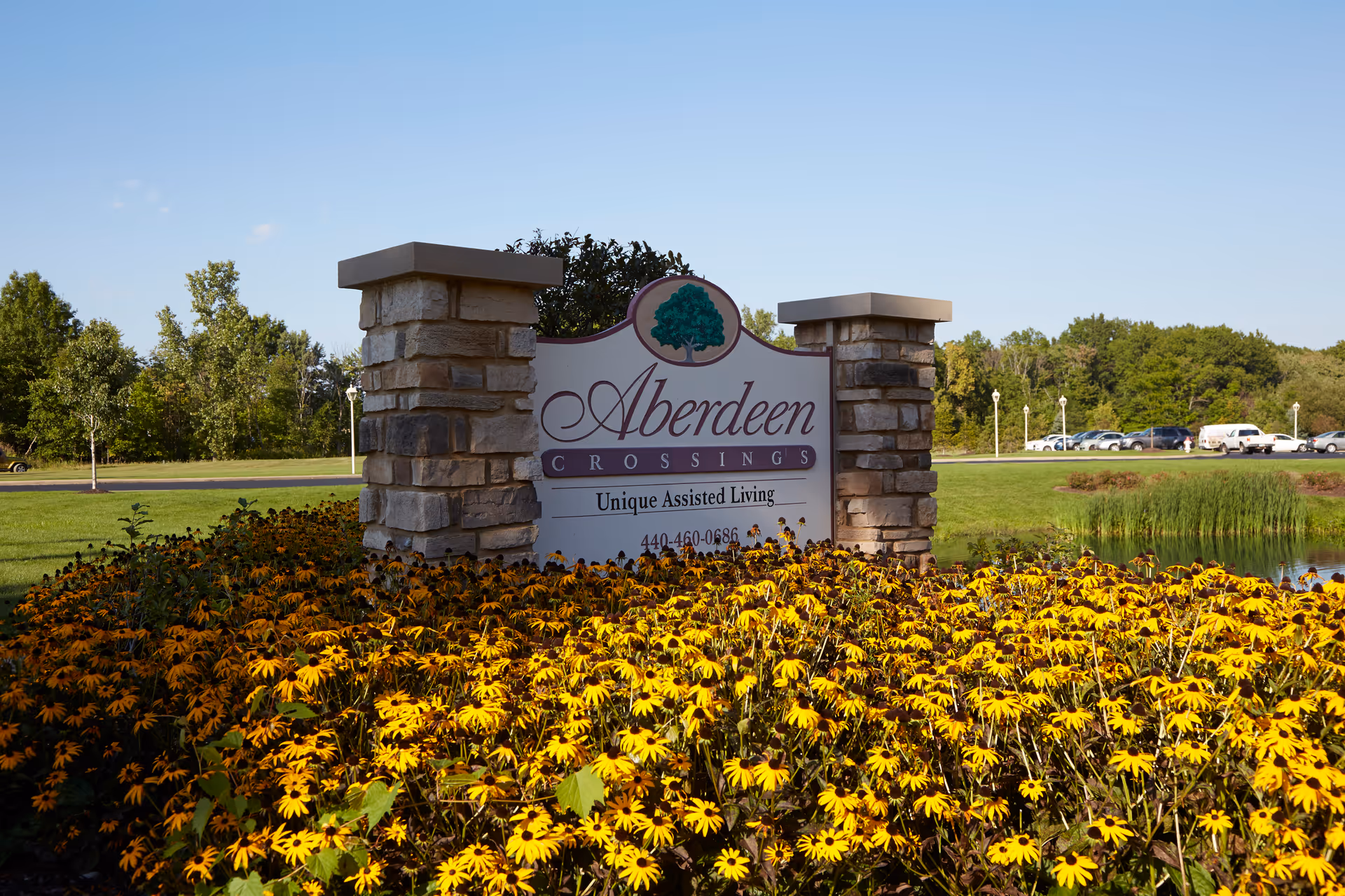 Entrance sign reading 'Aberdeen Crossings' for an assisted living community set between stone pillars amid yellow flowers and green lawn.