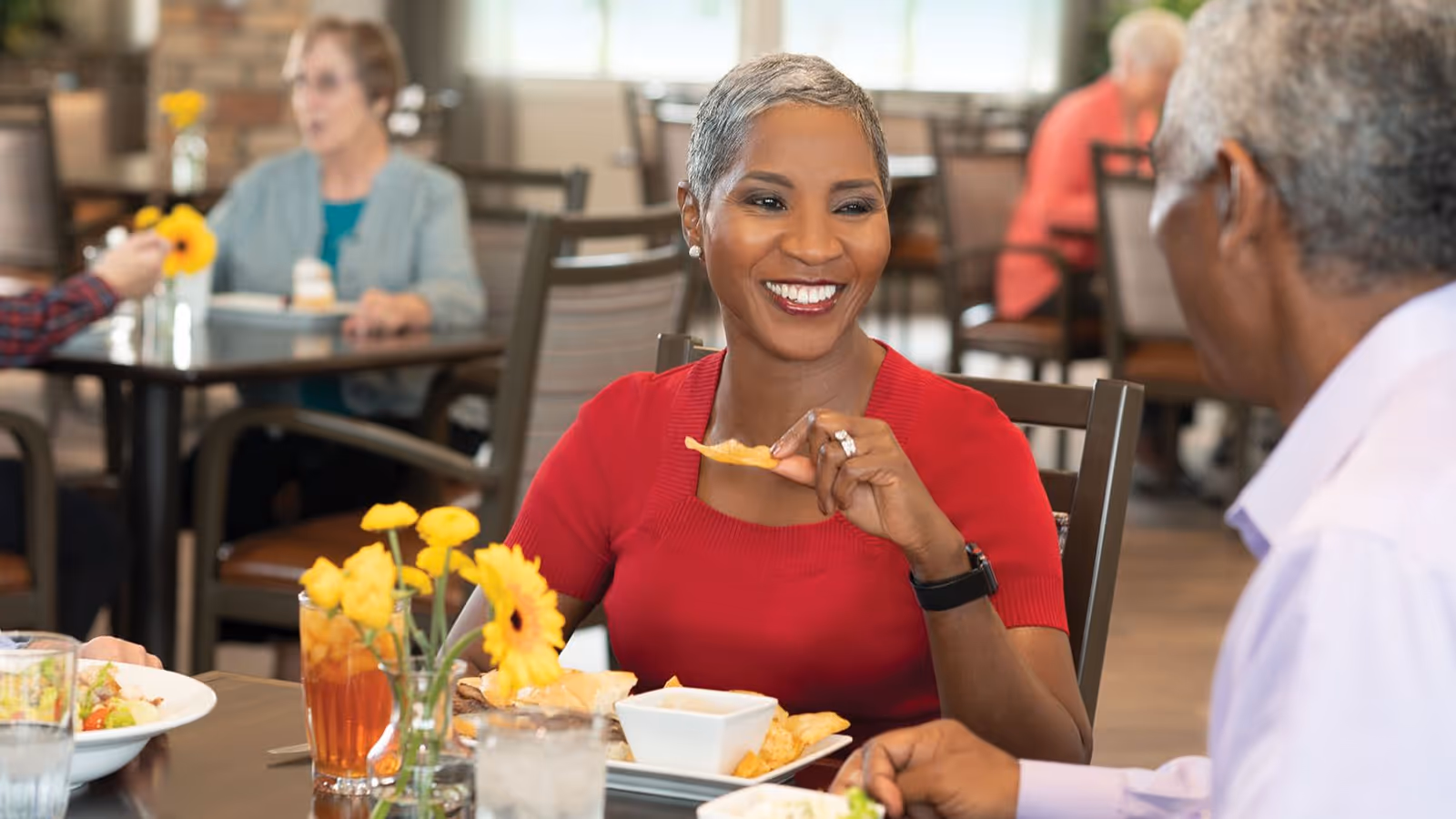A smiling woman in a red top is sitting at a dining table with a man, holding a chip and enjoying a meal together in a bright dining room with other people in the background.