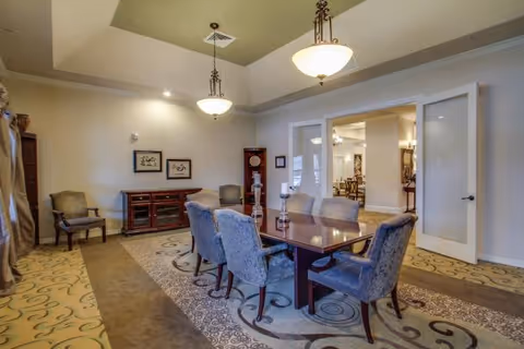 A formal dining room with a long wooden table surrounded by eight upholstered chairs. The room features a patterned carpet, two hanging light fixtures, a wooden sideboard against the wall, two framed artworks, and a grandfather clock. Double glass doors open to another room with additional seating visible.