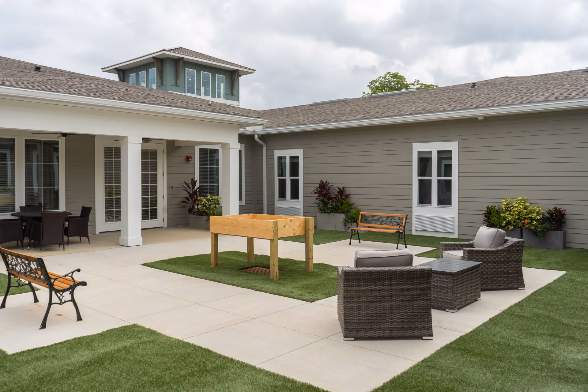 Outdoor courtyard area at The Canopy at Azalea Grove featuring patio furniture including a bench, two cushioned chairs, a small table, and a raised wooden planter box. The space is surrounded by a building with gray siding and white trim, with potted plants and a covered seating area with a table and chairs.